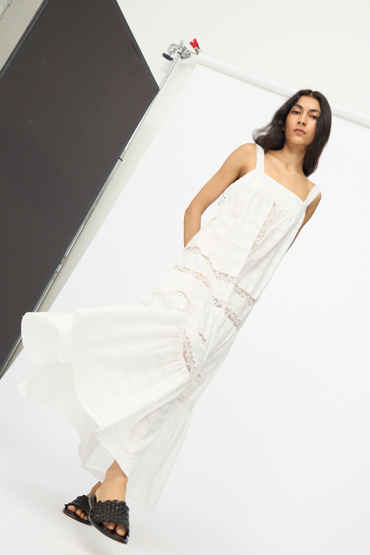 A woman wears the Loretta Caponi Populonia Dress in Off White, a long cotton maxi with lace details, paired with black woven sandals in a minimalist studio setting.