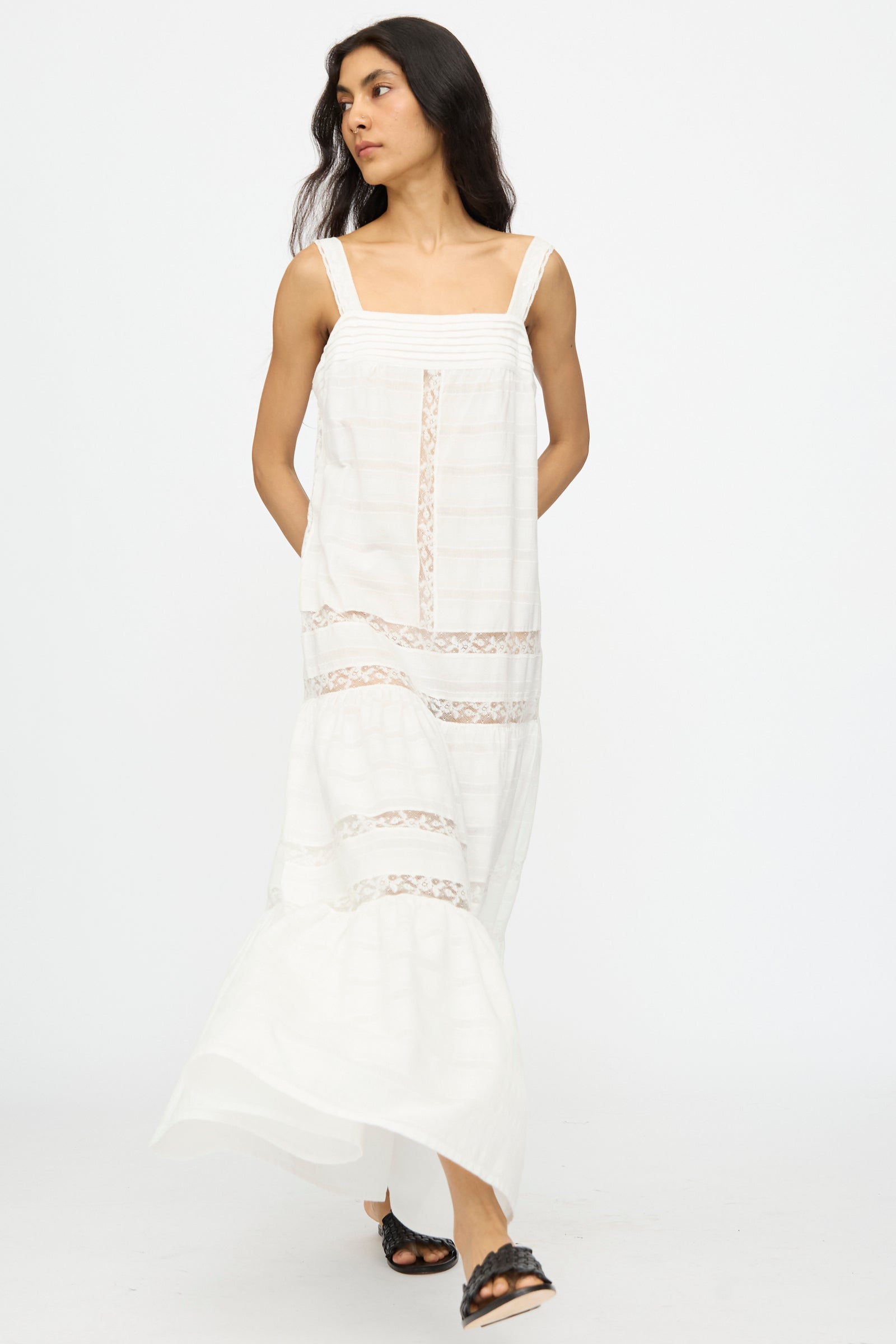 A woman wears the Loretta Caponi Populonia Dress in Off White—a long, sleeveless maxi with sheer lace details—paired with black sandals, posing against a plain white background.