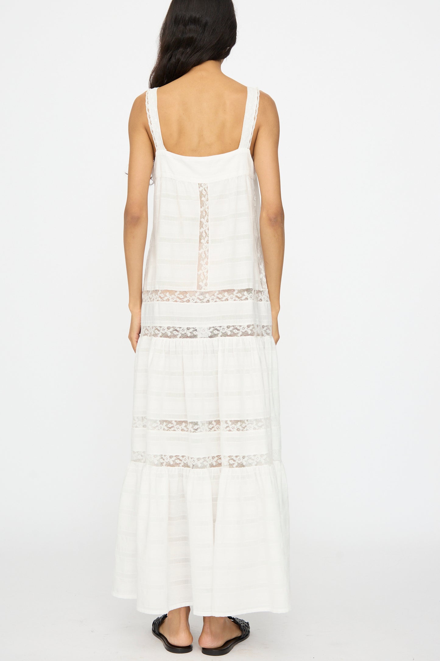 A woman with long dark hair stands facing away, wearing the Loretta Caponi Populonia Dress in Off White—a sleeveless cotton maxi with lace detailing—and black sandals, set against a plain white background.