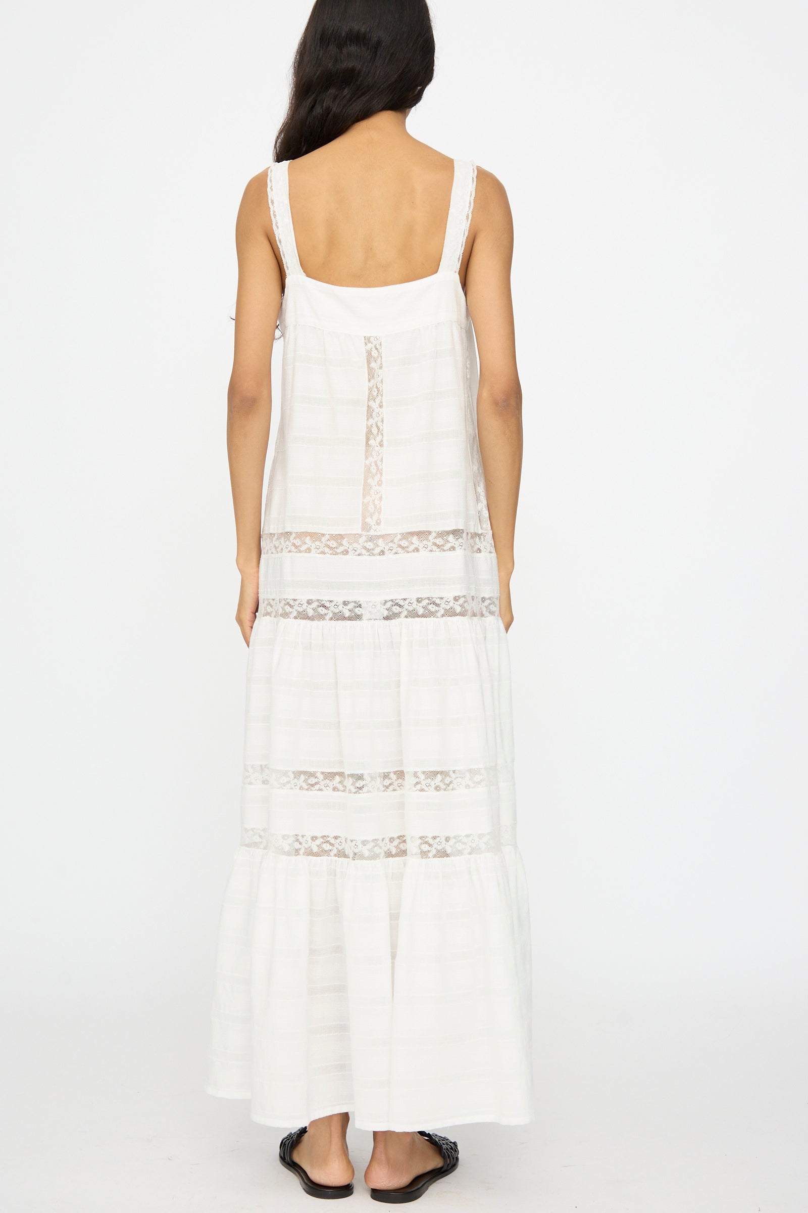 A woman with long dark hair stands facing away, wearing the Loretta Caponi Populonia Dress in Off White—a sleeveless cotton maxi with lace detailing—and black sandals, set against a plain white background.