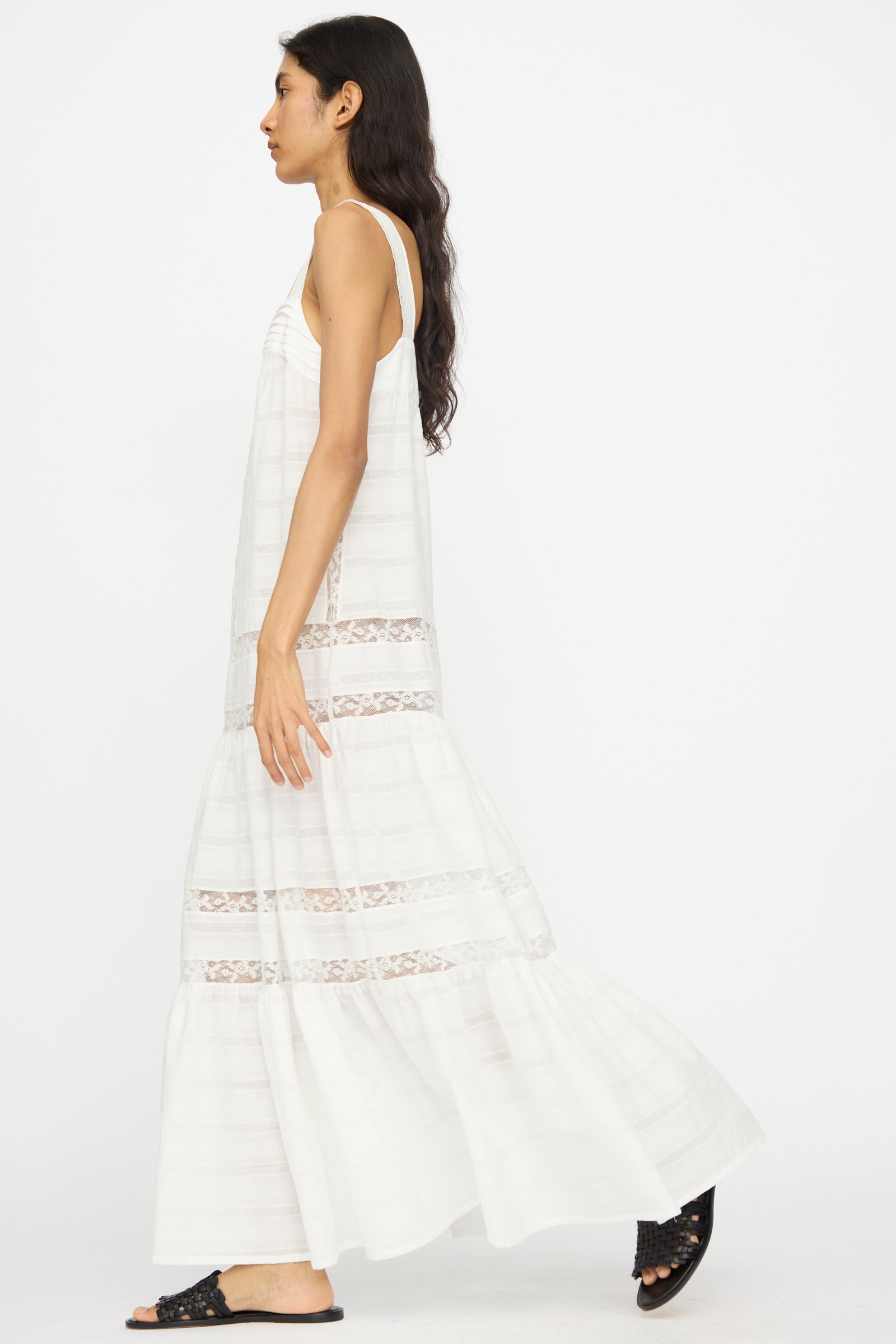 A woman in the Loretta Caponi Populonia Dress in Off White, a sleeveless cotton maxi with lace details, walks left wearing black slide sandals against a plain white background.