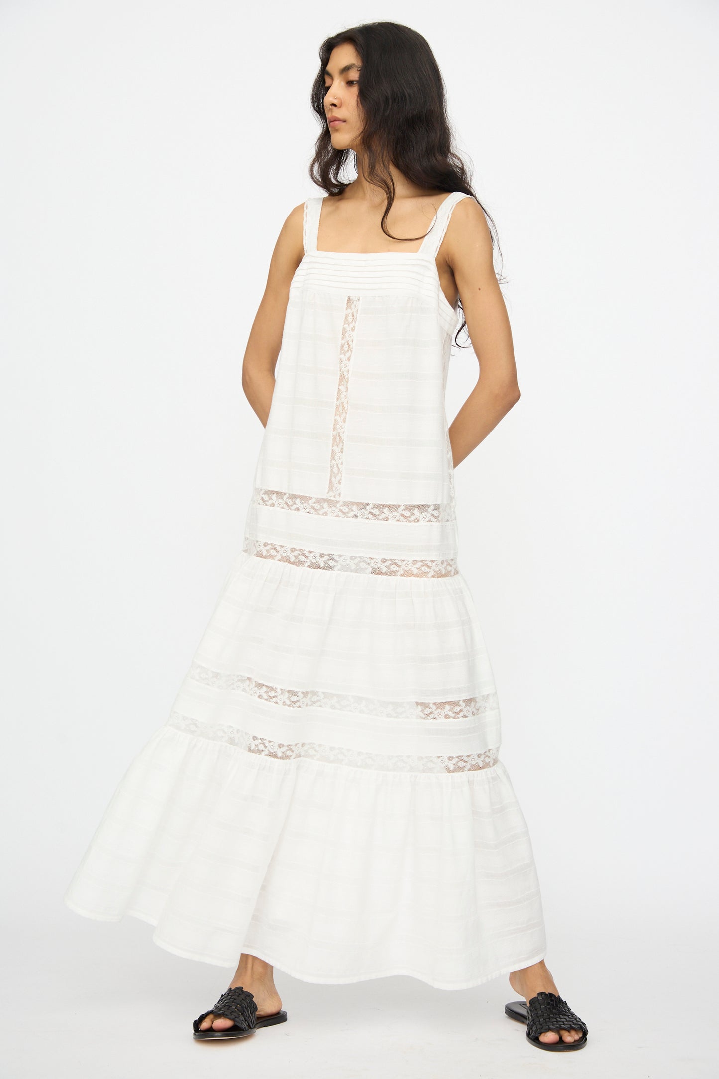 A woman with long dark hair wears the Loretta Caponi Populonia Dress in Off White—an ankle-length, sleeveless cotton maxi with lace detailing—paired with black sandals, standing against a plain white background.