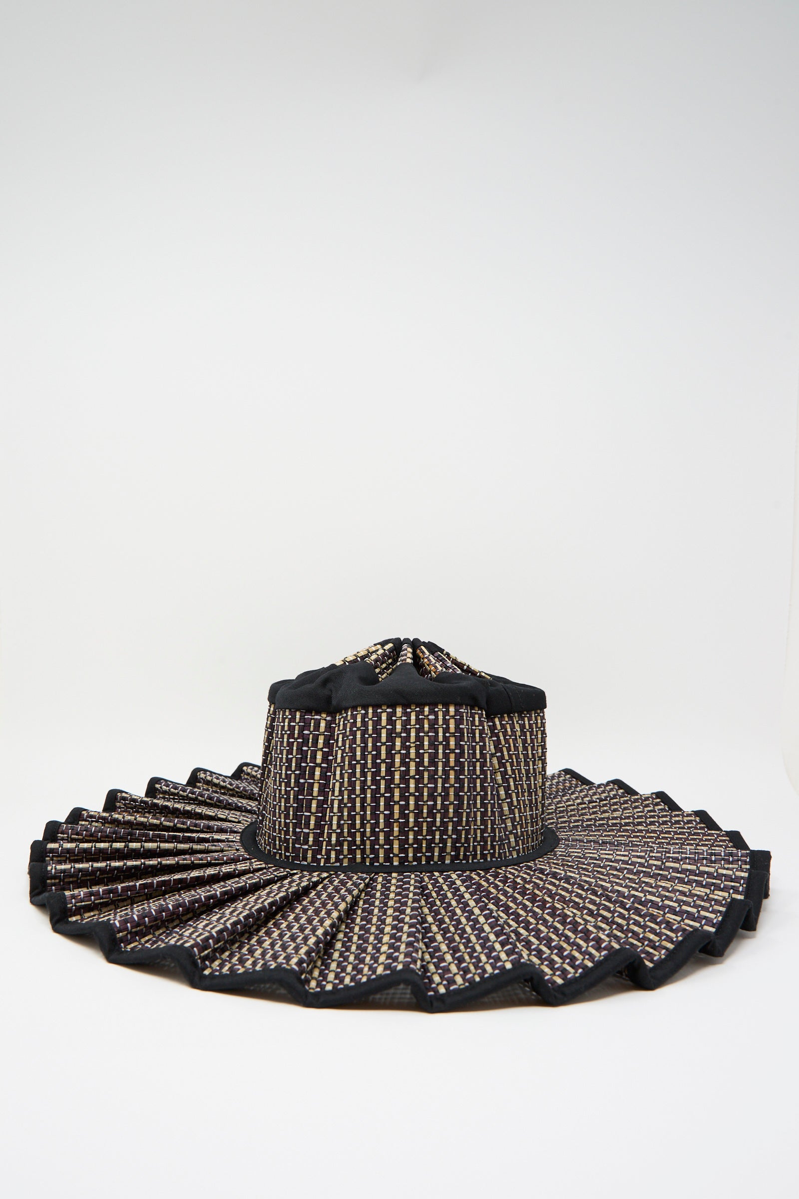 The Lorna Murray Melbourne Island Capri Maxi Hat is a wide-brimmed sun hat woven from natural grass fibers, featuring a black, beige, and brown checkered pattern with a pleated brim and UPF 50+ sun protection, displayed on a white background.