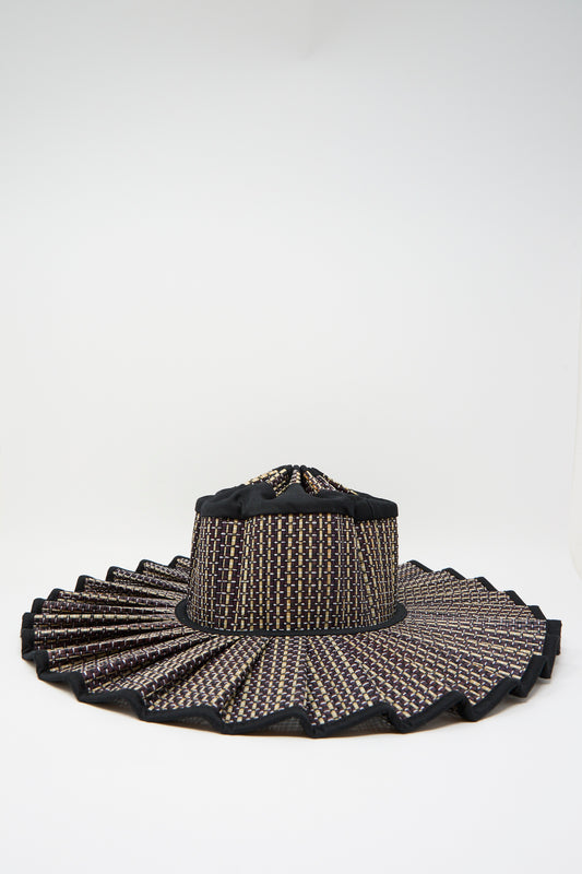 The Lorna Murray Melbourne Island Capri Maxi Hat is a wide-brimmed sun hat woven from natural grass fibers, featuring a black, beige, and brown checkered pattern with a pleated brim and UPF 50+ sun protection, displayed on a white background.
