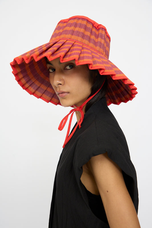 Wearing the Island Capri Hat in Bossa Nova by Lorna Murray—a wide-brimmed, red and orange plaid sun hat with ruffled edges—this person poses in a black sleeveless top against a plain white background.