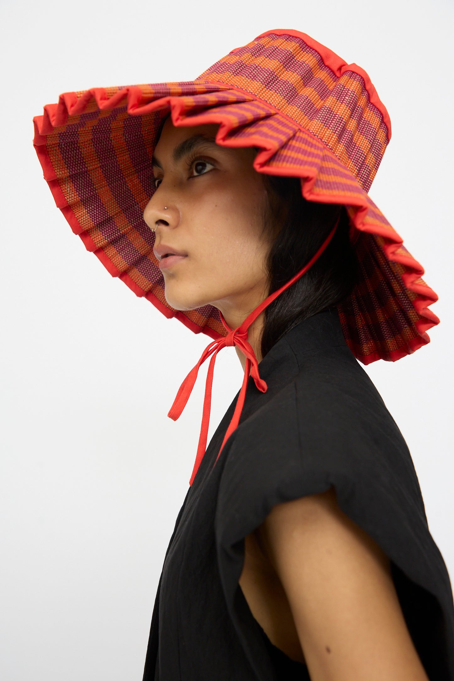 A person in a black top wears the Lorna Murray Island Capri Hat in Bossa Nova—a wide-brimmed, red and orange plaid sun hat with ruffled edges and chin tie—shown in profile; crafted for style and UPF 50+ protection.