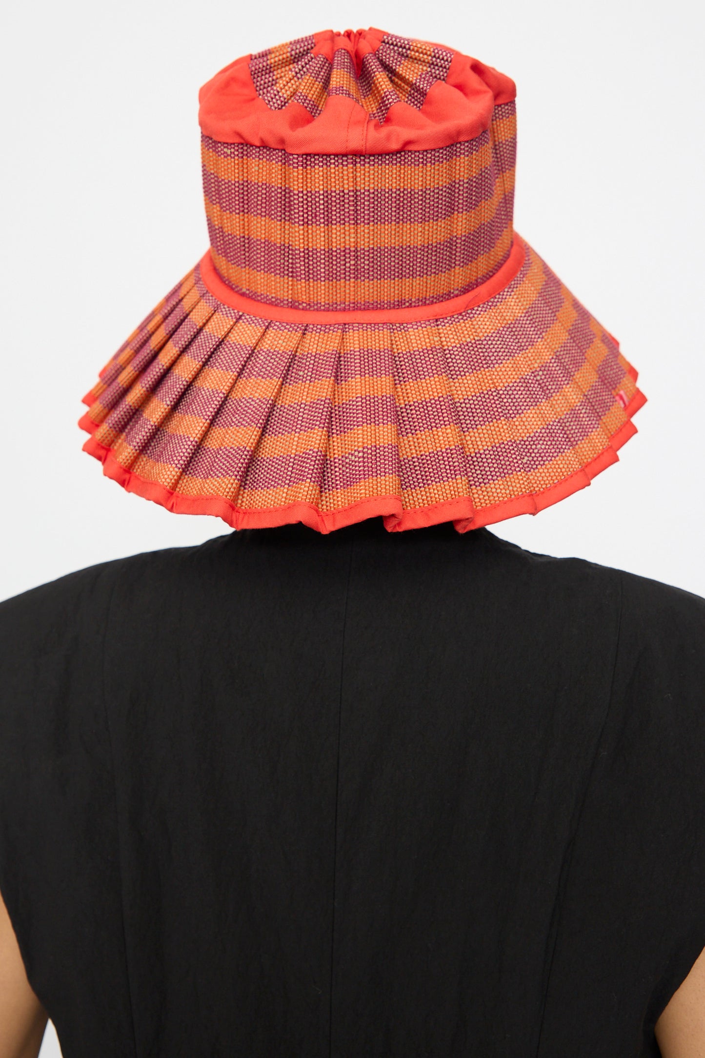 A person shown from the back wears Lorna Murray's Island Capri Hat in Bossa Nova—an orange and purple checkered sun hat made from natural grass fibers—with a black top, set against a plain background.
