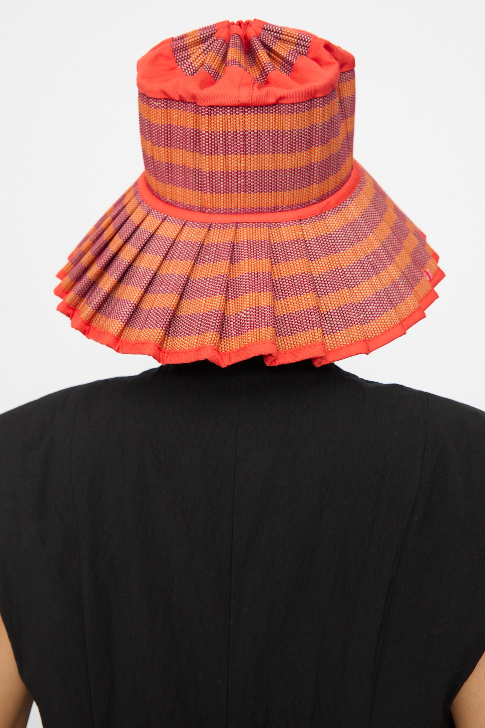 A person shown from the back wears Lorna Murray's Island Capri Hat in Bossa Nova—an orange and purple checkered sun hat made from natural grass fibers—with a black top, set against a plain background.