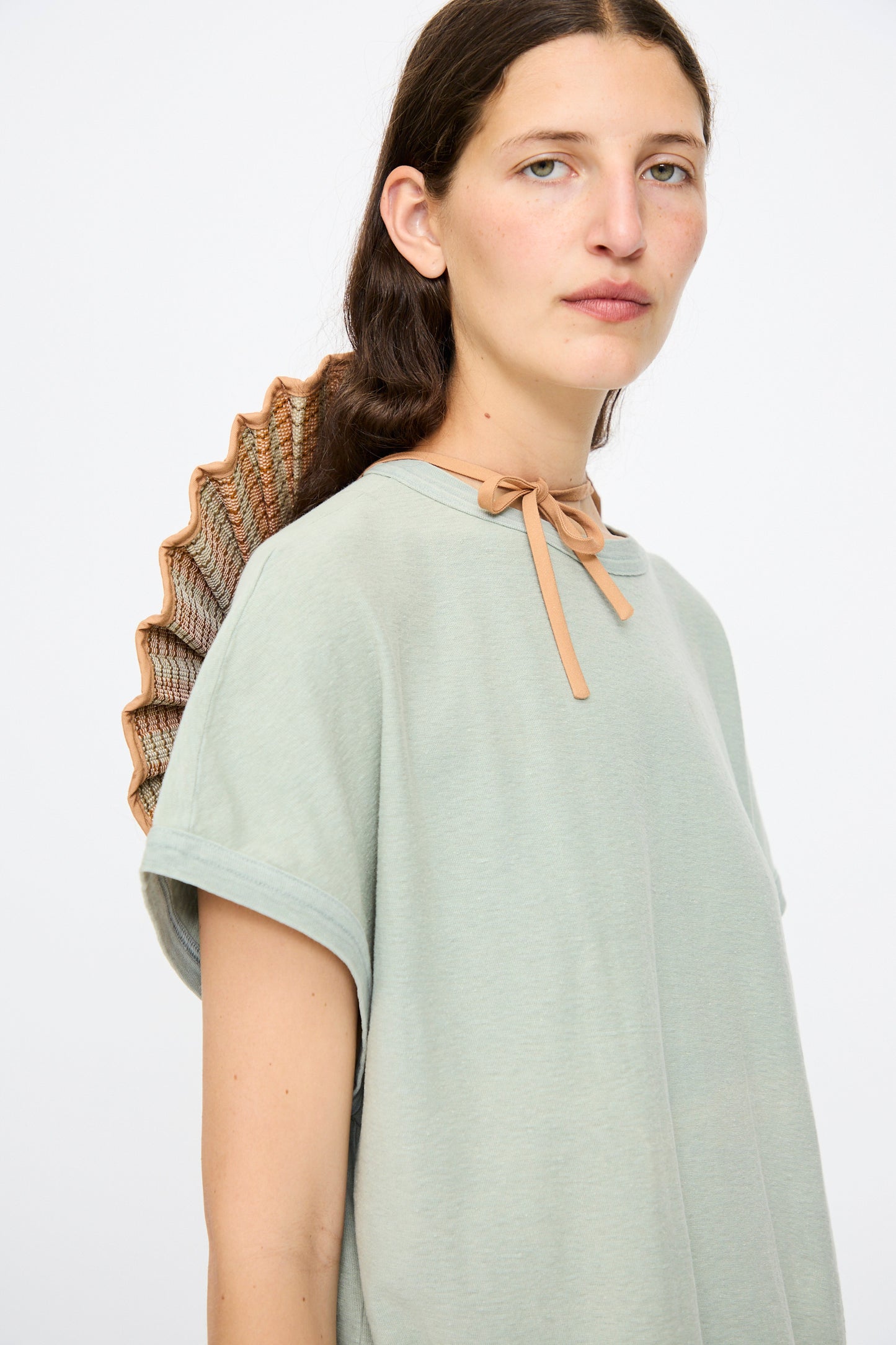 A woman wears the Lorna Murray Island Capri Maxi Hat in Petra, a pleated, fan-shaped accessory made of natural grass fibers and tied with a tan ribbon, paired with a loose, light green short-sleeve shirt.