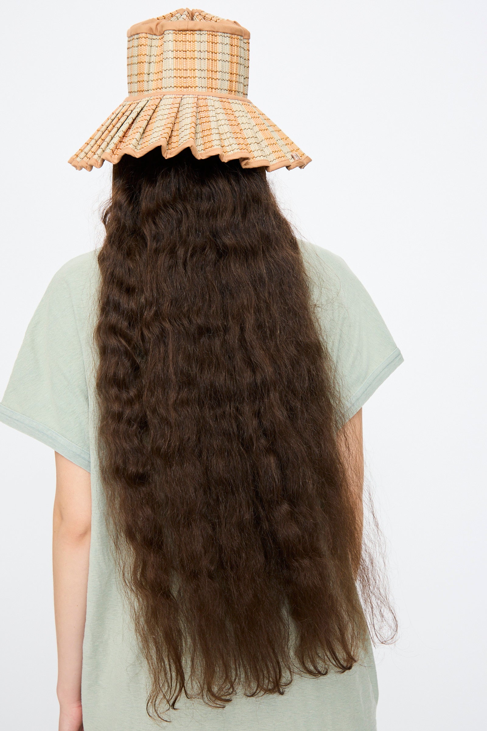 Person with long, wavy brown hair wears the Lorna Murray Island Capri Maxi Hat in Petra—a pleated, checked tan sun hat—paired with a light green shirt, photographed from behind against a plain background.