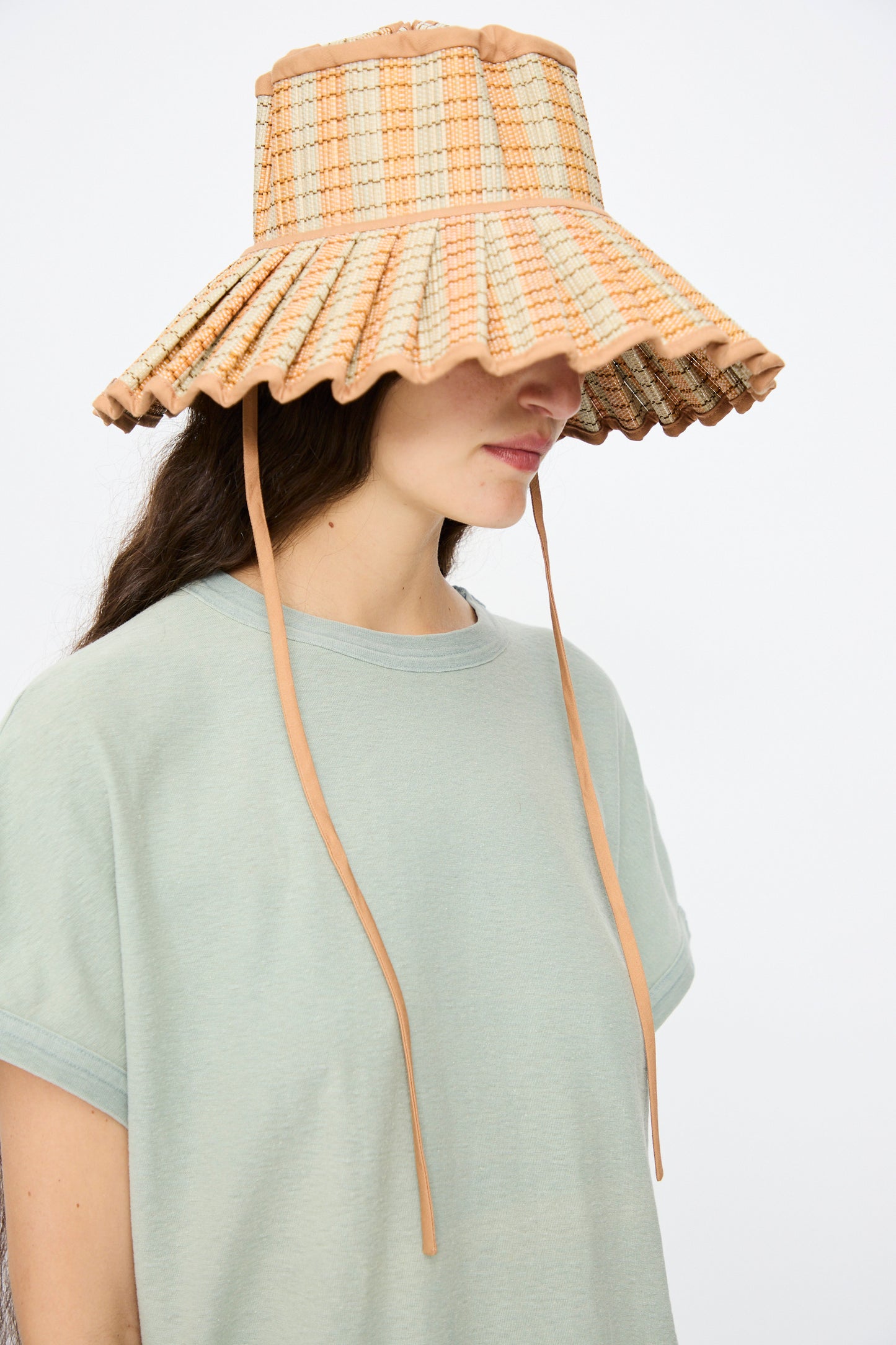 A person wearing a light green t-shirt and the Lorna Murray Island Capri Maxi Hat in Petra, featuring plaid patterns, long chin straps, and UPF 50+ sun protection, stands against a plain background.