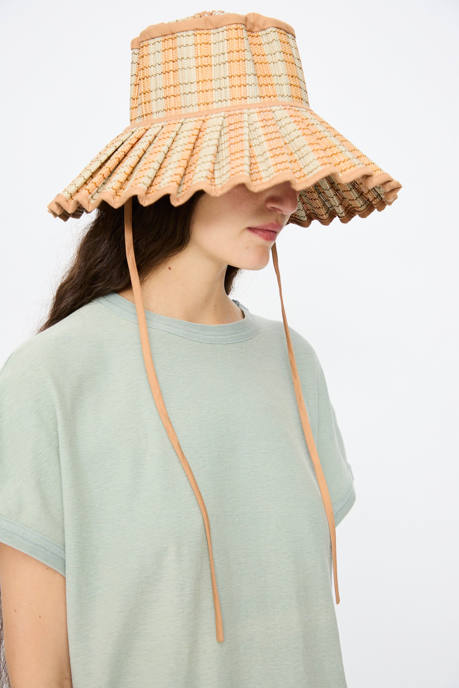 A person wearing a light green t-shirt and the Lorna Murray Island Capri Maxi Hat in Petra, featuring plaid patterns, long chin straps, and UPF 50+ sun protection, stands against a plain background.