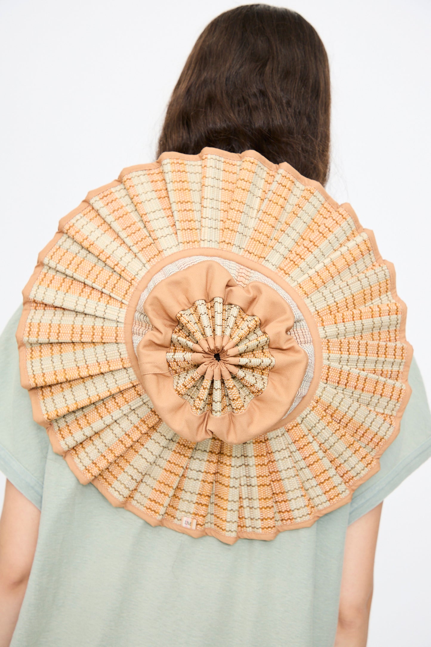 A person with long brown hair is seen from behind wearing the Lorna Murray Island Capri Maxi Hat in Petra, a large round pleated sun hat in beige and light brown, made from natural grass fibers with UPF 50+ protection.