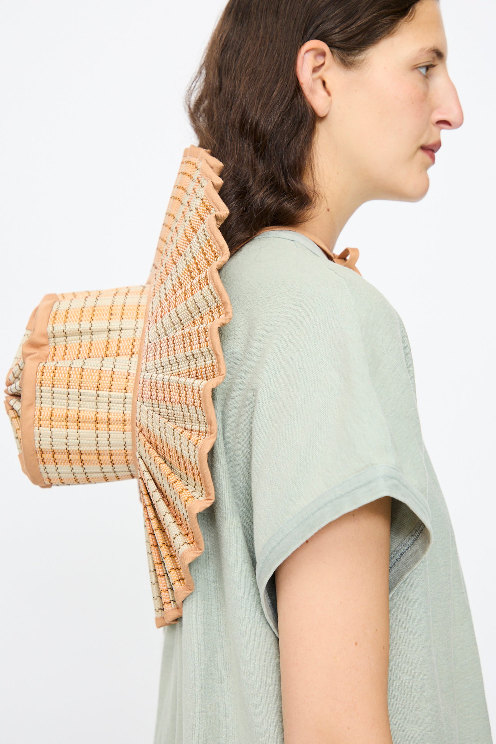 A person in a light green shirt wears the Lorna Murray Island Capri Maxi Hat in Petra, featuring a wide, ruffled brim made from natural grass fibers, on their back while facing sideways against a plain background.