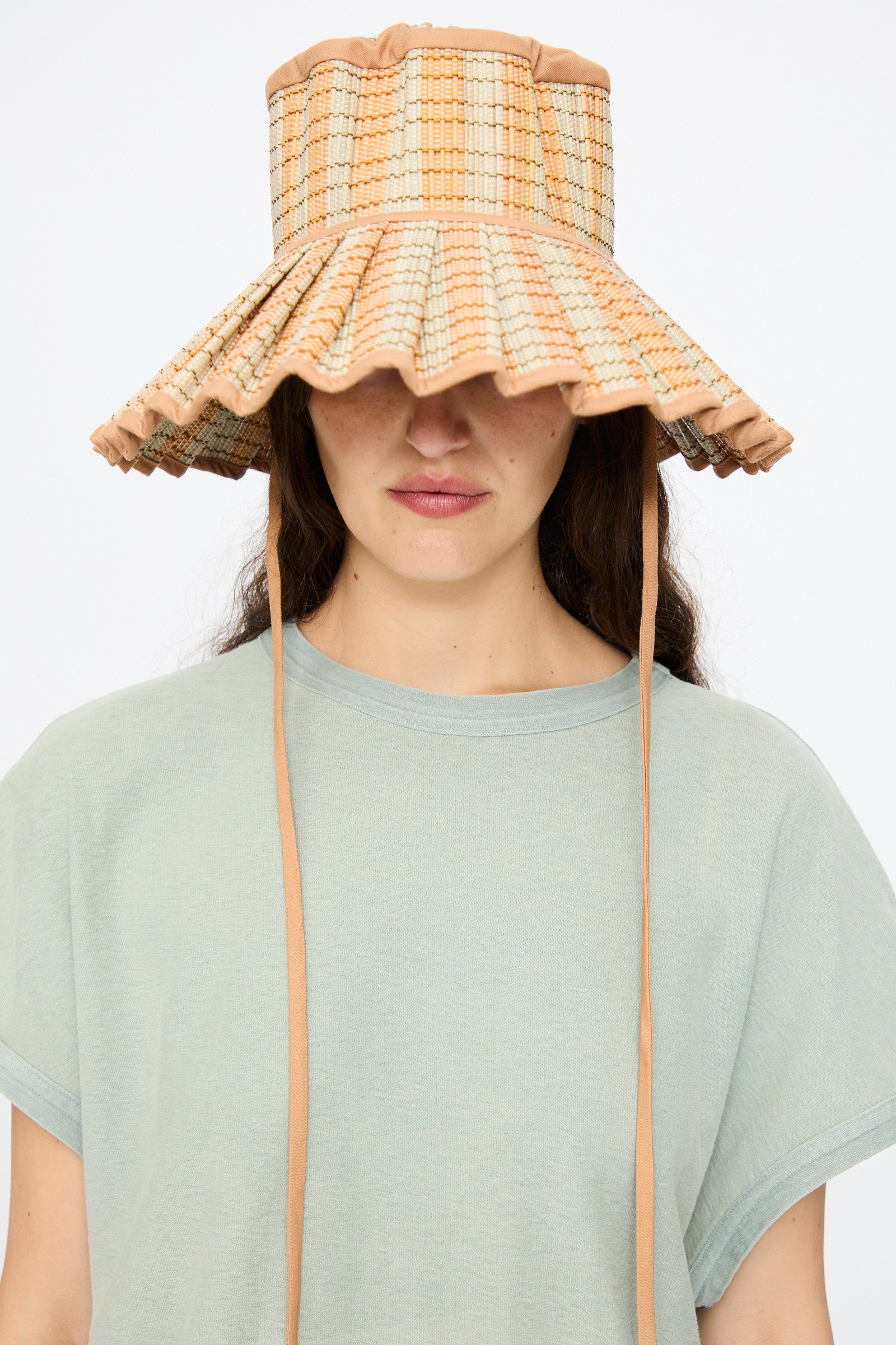 A woman in a light green t-shirt faces the camera against a plain background, wearing the Lorna Murray Island Capri Maxi Hat in Petra—a wide-brimmed, checkered sun hat crafted from natural grass fibers with chin straps.