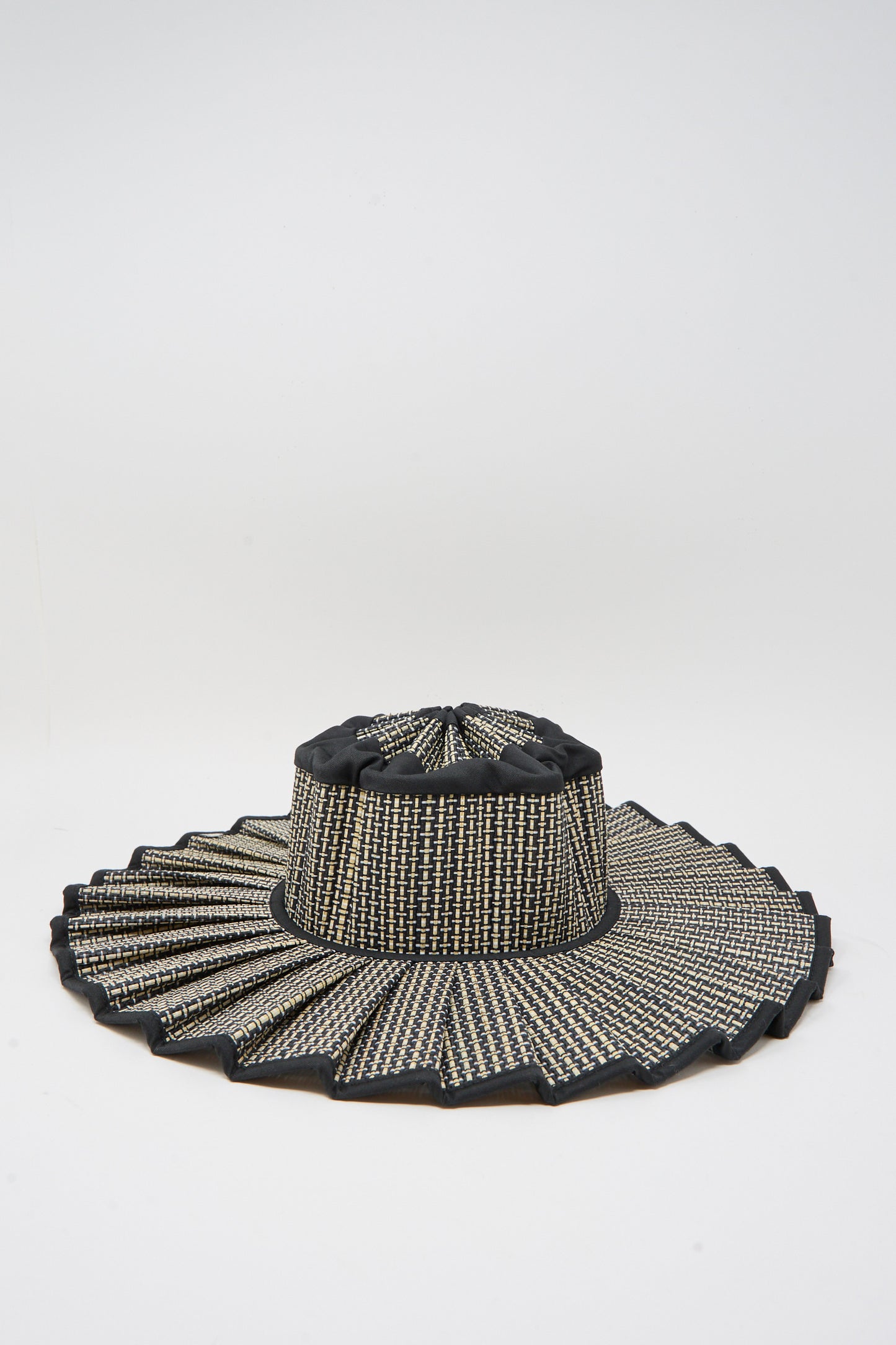 The Island Capri Maxi Hat in Melbourne by Lorna Murray is a wide-brim sun hat woven from natural grass fibers with a pleated zigzag edge, black and beige checkered pattern, UPF 50+ protection, shown on a plain white background.