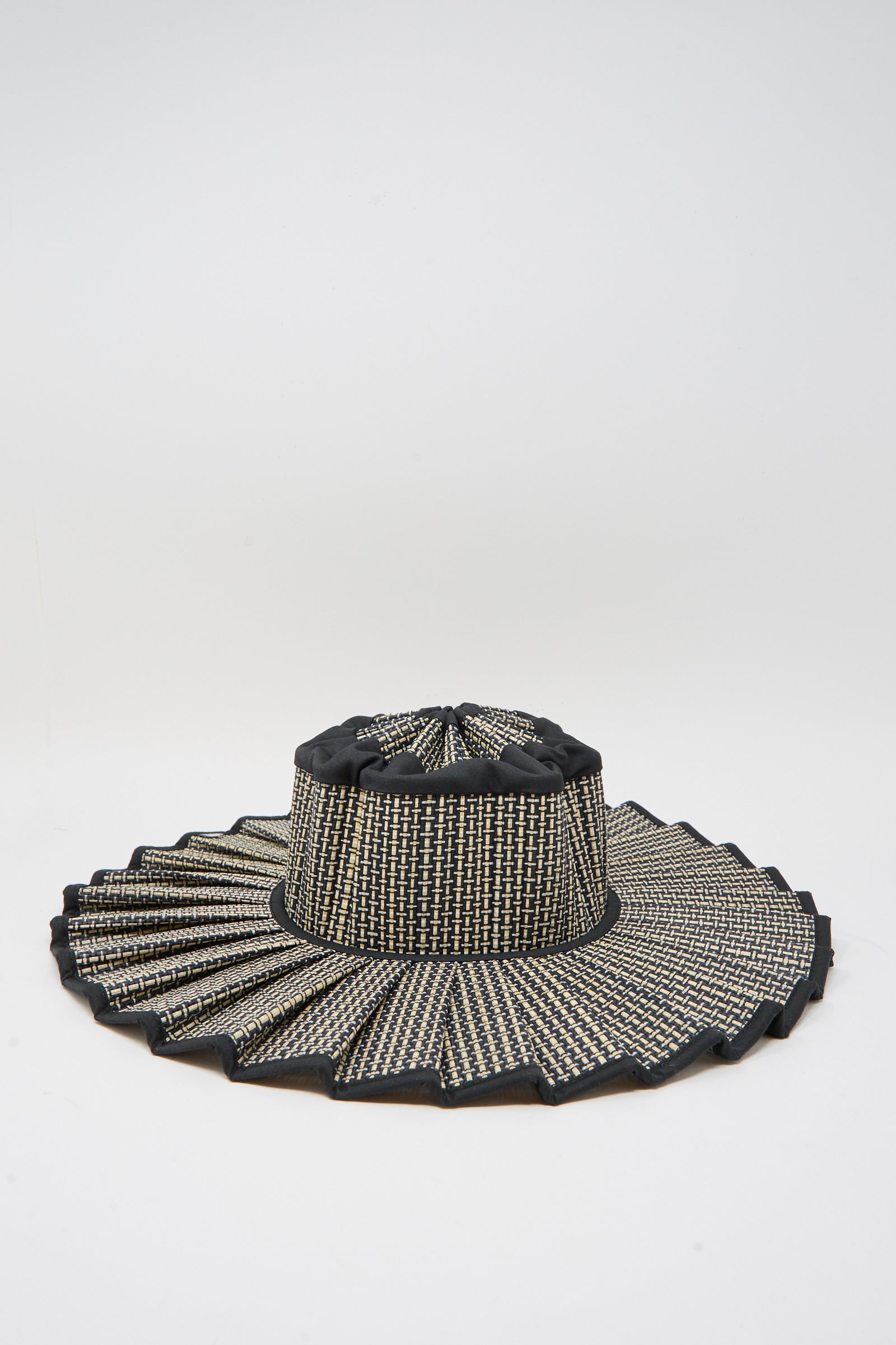 The Island Capri Maxi Hat in Melbourne by Lorna Murray is a wide-brim sun hat woven from natural grass fibers with a pleated zigzag edge, black and beige checkered pattern, UPF 50+ protection, shown on a plain white background.