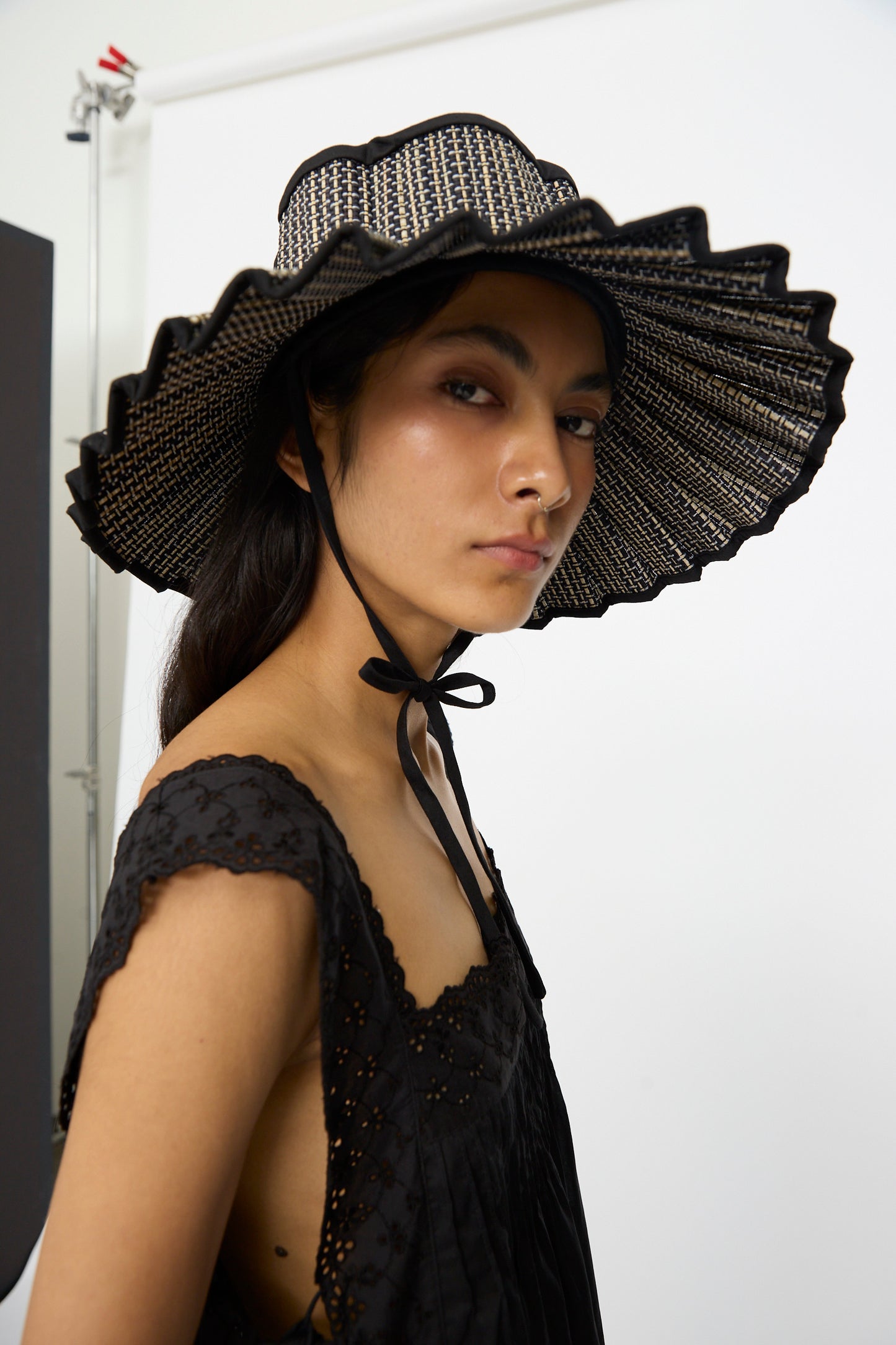 A person wears the Lorna Murray Island Capri Maxi Hat in Melbourne, featuring a wide brim of natural grass fibers with wavy edges and a black sleeveless top, standing against a plain white background. The hat provides stylish UPF 50+ protection.