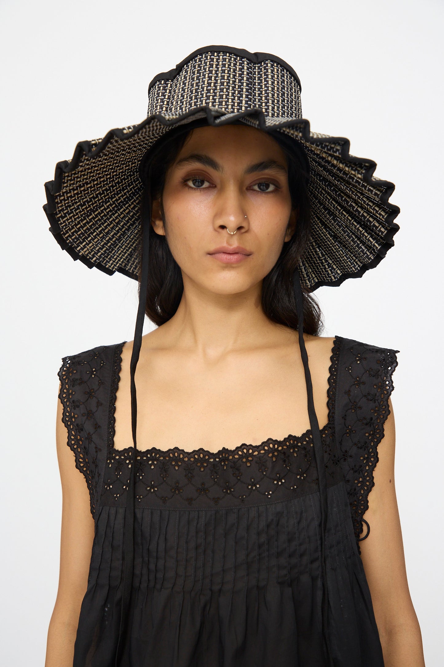 A woman stands against a plain white background wearing the Lorna Murray Island Capri Maxi Hat in Melbourne, a UPF 50+ sun hat crafted from natural grass fibers with black trim, paired with a black sleeveless eyelet top.