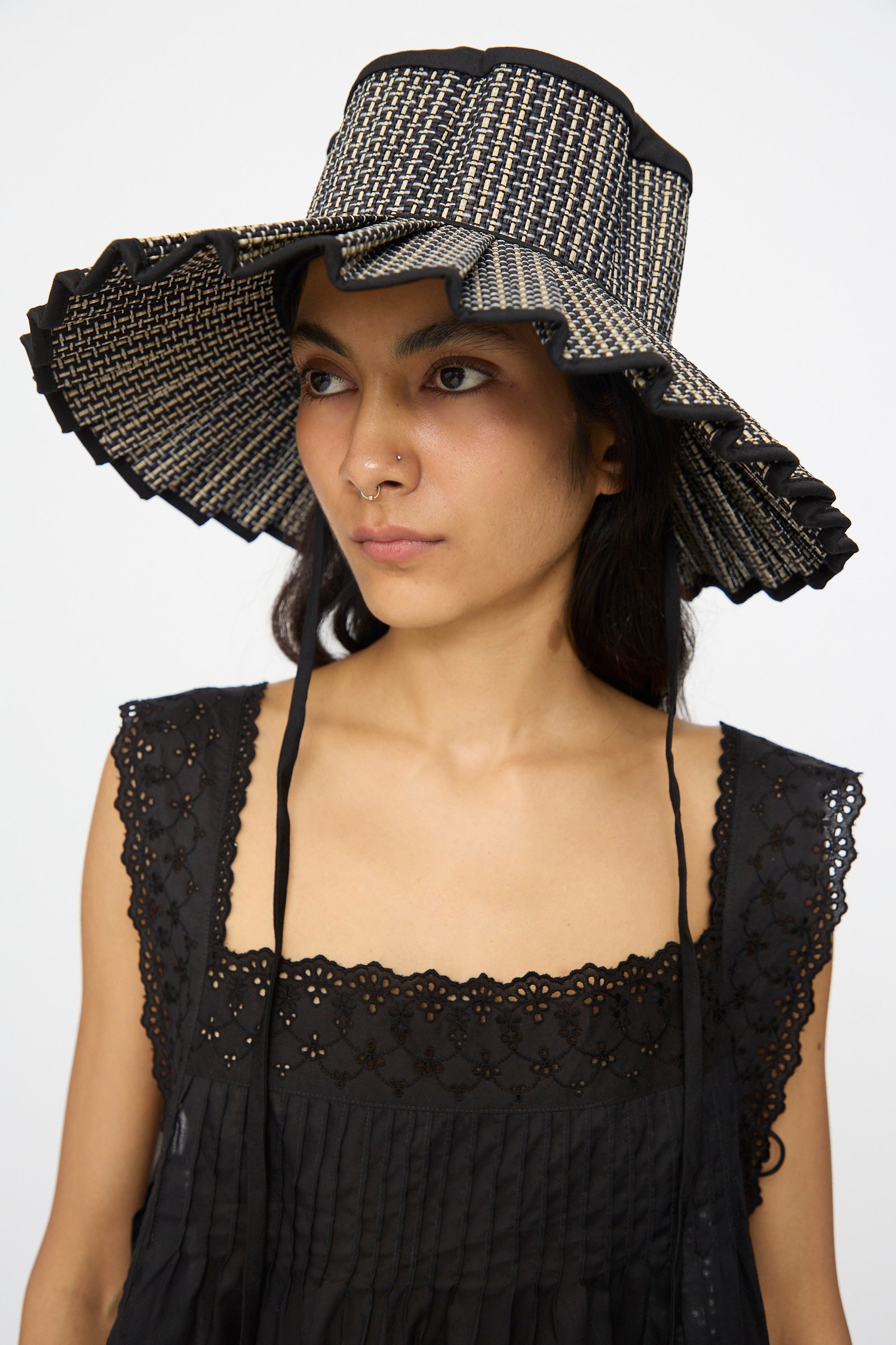 A woman wearing the Lorna Murray Island Capri Maxi Hat in Melbourne, a wide-brimmed sun hat with black and beige checks and ruffled edges, paired with a black lace-trimmed dress, gazes slightly to the side.