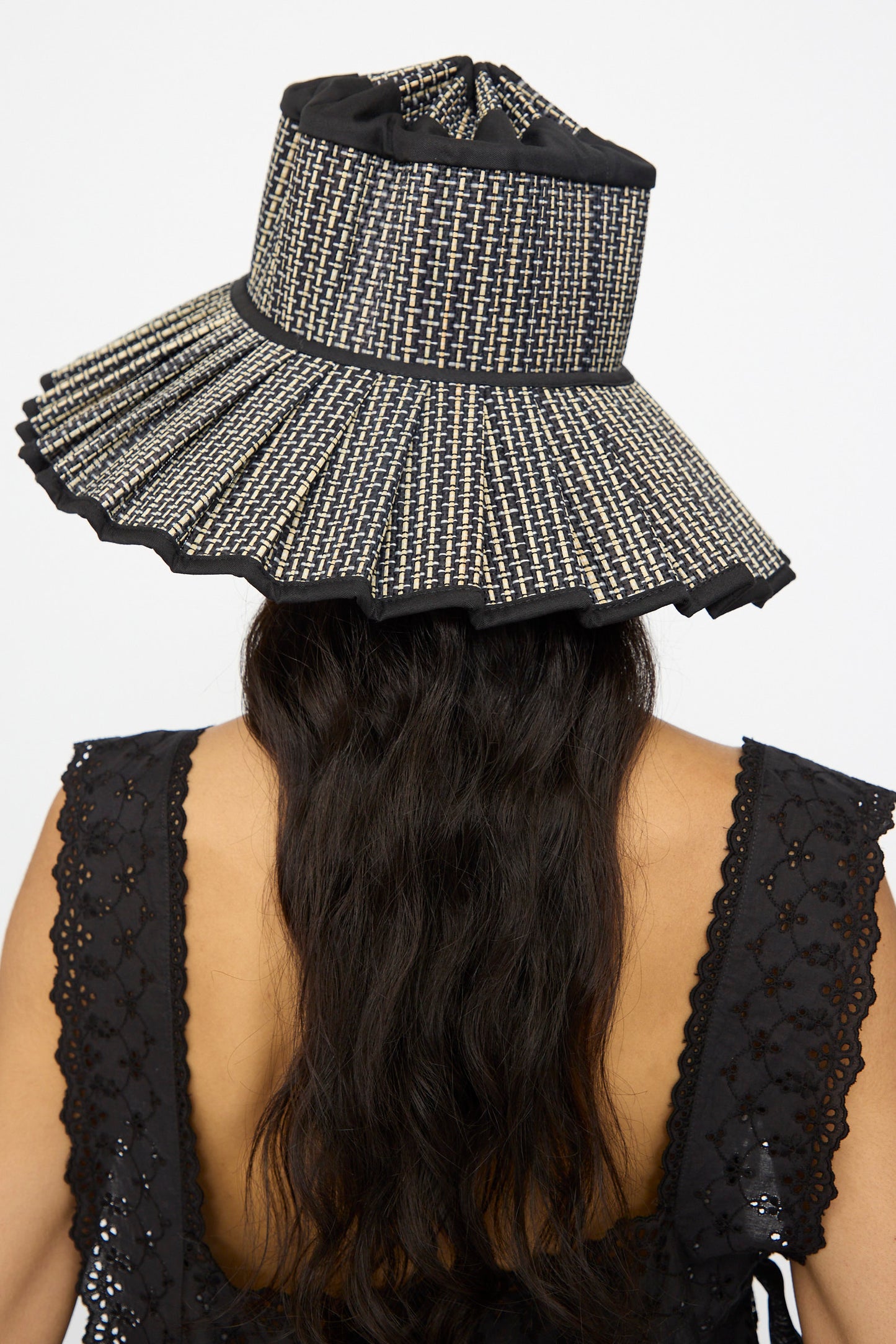 A person with long wavy hair wears the Lorna Murray Island Capri Maxi Hat in Melbourne, a wide-brimmed sun hat woven from natural grass fibers, paired with a sleeveless black eyelet top, seen from behind.