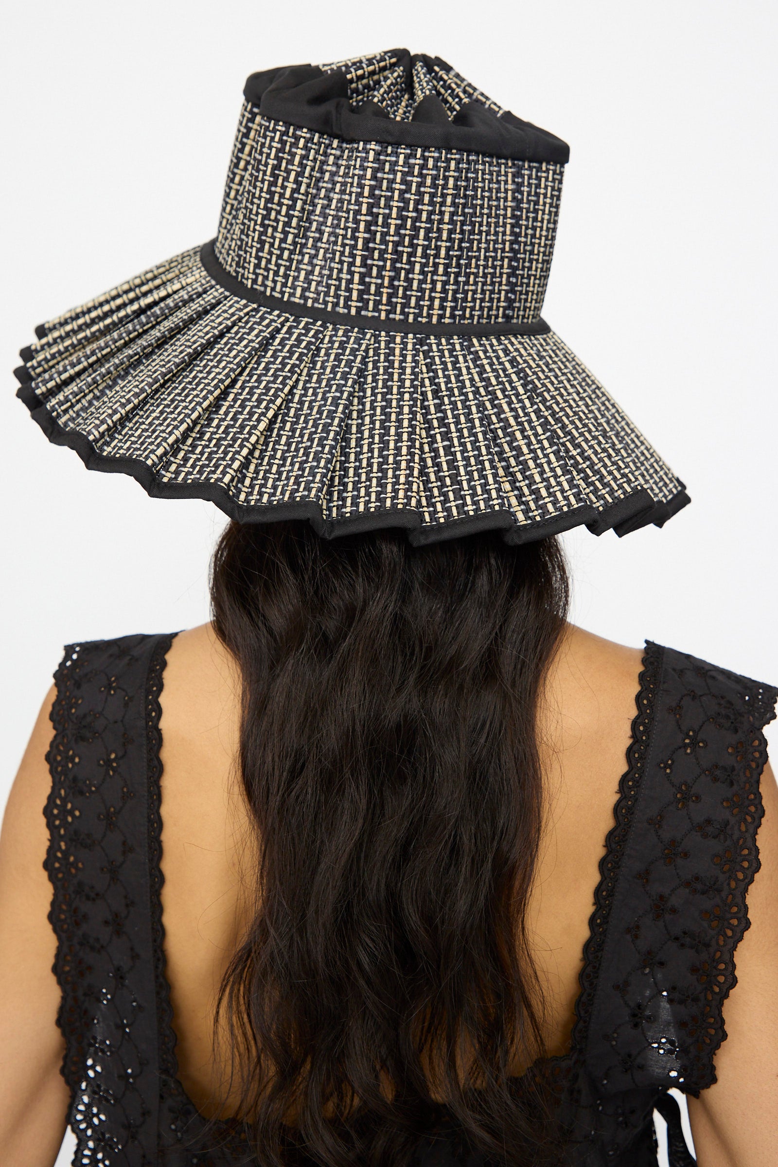 A person with long wavy hair wears the Lorna Murray Island Capri Maxi Hat in Melbourne, a wide-brimmed sun hat woven from natural grass fibers, paired with a sleeveless black eyelet top, seen from behind.