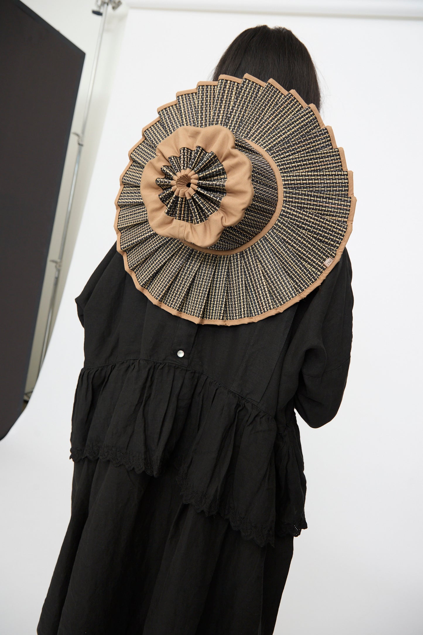 A person in black faces away from the camera, displaying the Lorna Murray Island Capri Maxi Hat in Nara—an oversized, pleated sun hat crafted from natural grass fibers in tan and black—against a white background.