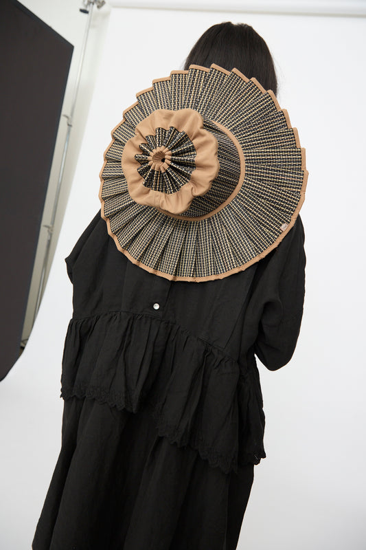 A person in black faces away from the camera, displaying the Lorna Murray Island Capri Maxi Hat in Nara—an oversized, pleated sun hat crafted from natural grass fibers in tan and black—against a white background.