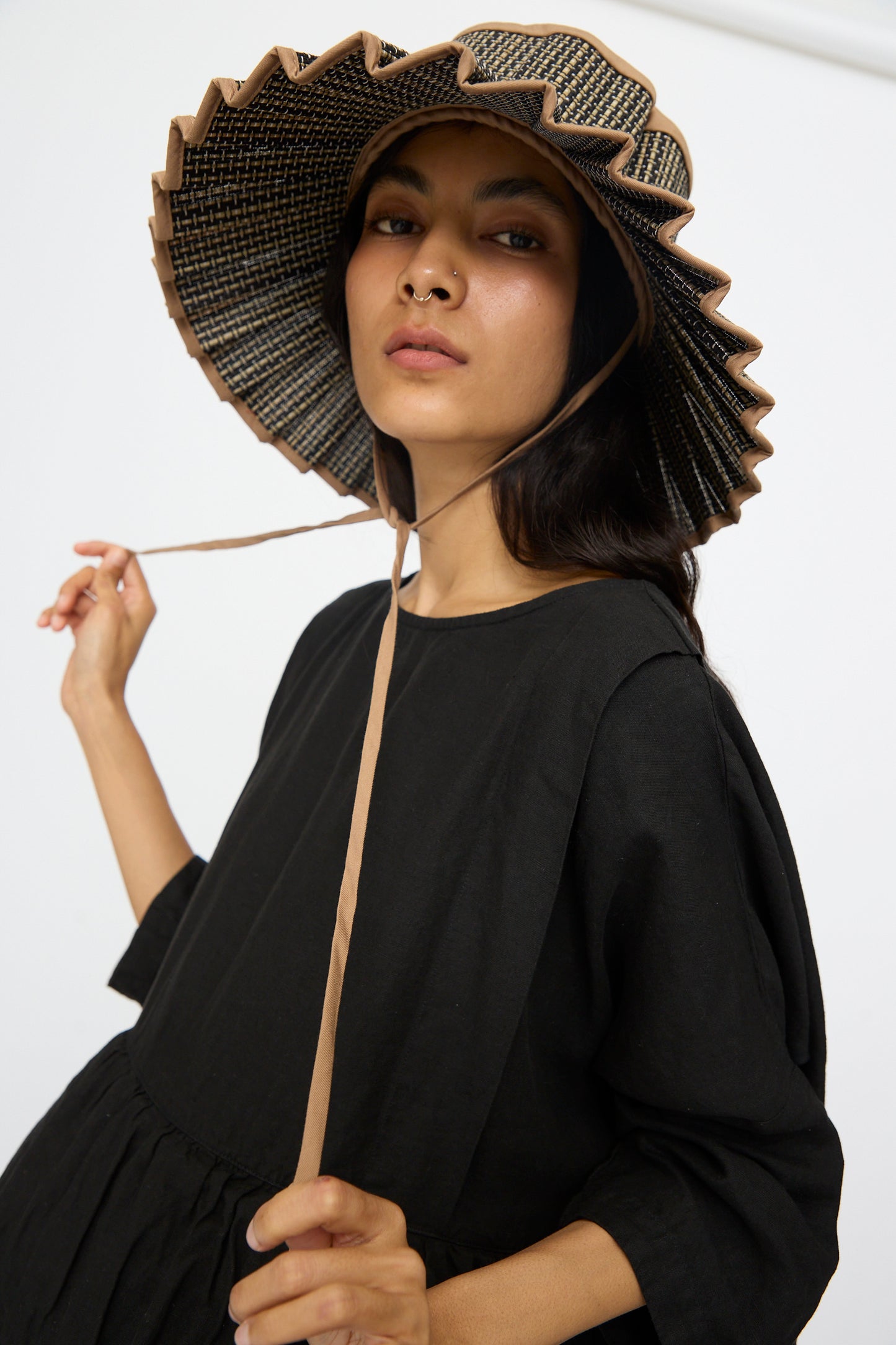 A person wears the Lorna Murray Island Capri Maxi Hat in Nara, made of natural grass fibers, paired with a loose black dress and looking at the camera while holding the hat’s strap.