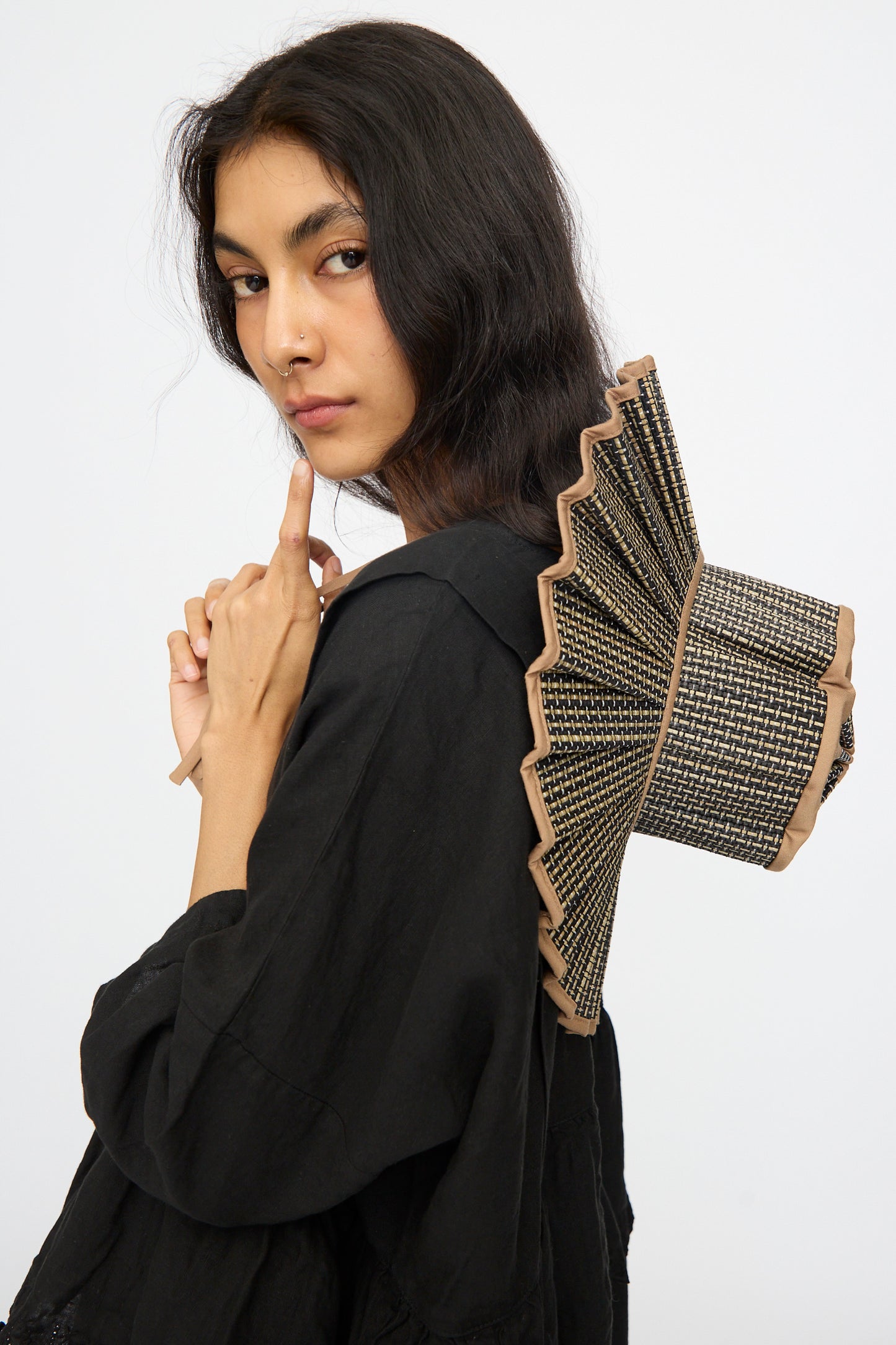 A woman in a black outfit poses with her finger near her lips, wearing the Lorna Murray Island Capri Maxi Hat in Nara, its fan-shaped brim of natural grass fibers tilted stylishly atop her head against a plain white background.