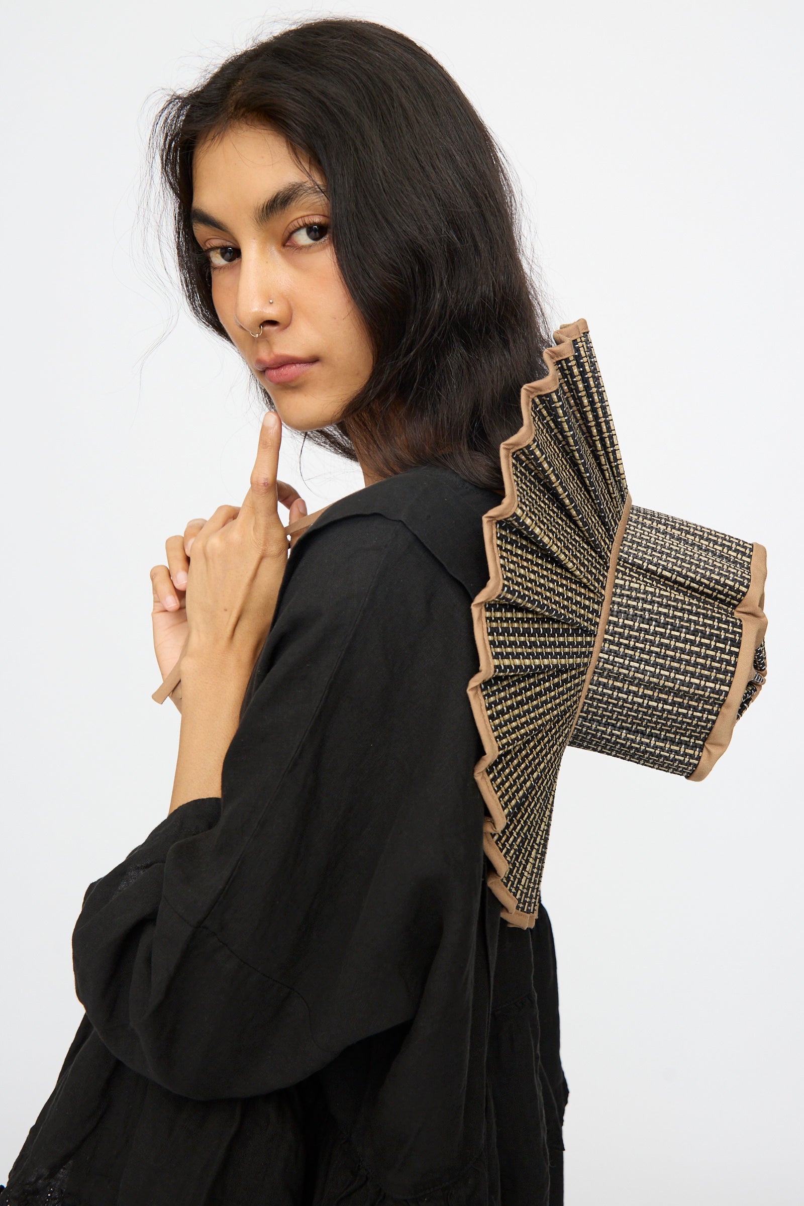 A woman in a black outfit poses with her finger near her lips, wearing the Lorna Murray Island Capri Maxi Hat in Nara, its fan-shaped brim of natural grass fibers tilted stylishly atop her head against a plain white background.