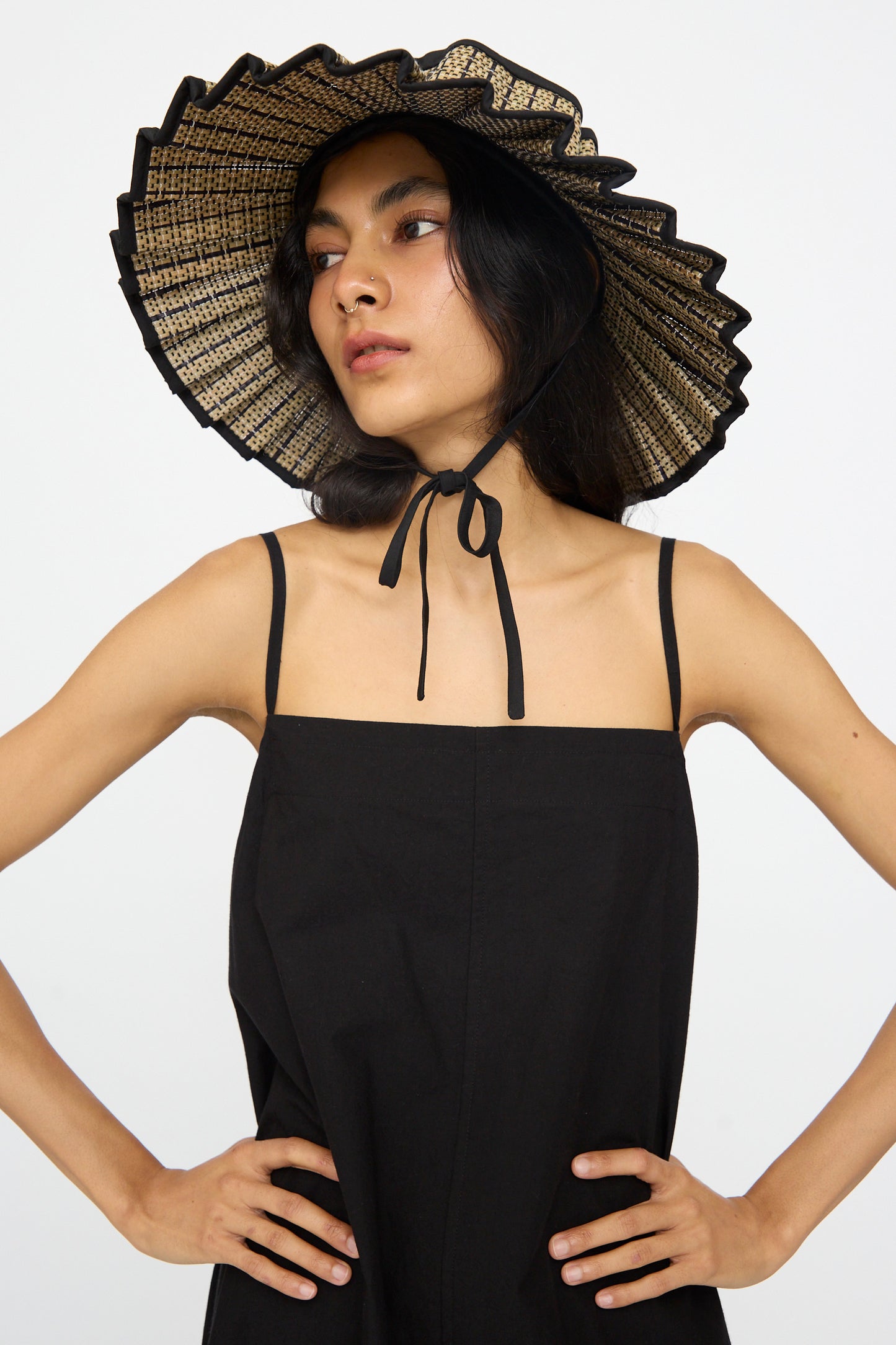 A woman in a black spaghetti-strap dress looks to the side against a plain background, wearing the Lorna Murray Island Capri Maxi Hat in Lovers Point—crafted from natural grass fibers with a ruffled edge and UPF 50+ protection.