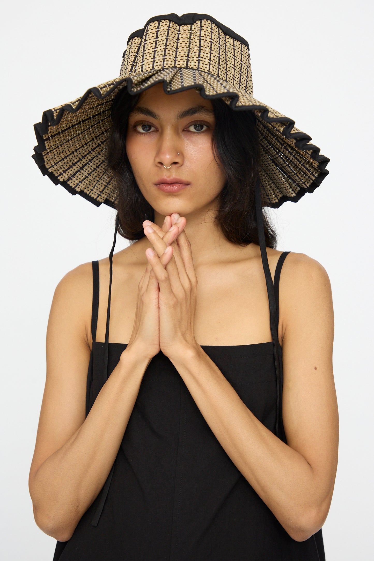 A woman in the Island Capri Maxi Hat in Lovers Point by Lorna Murray, made from natural grass fibers, and a black sleeveless dress stands with her hands clasped, looking at the camera against a plain background.