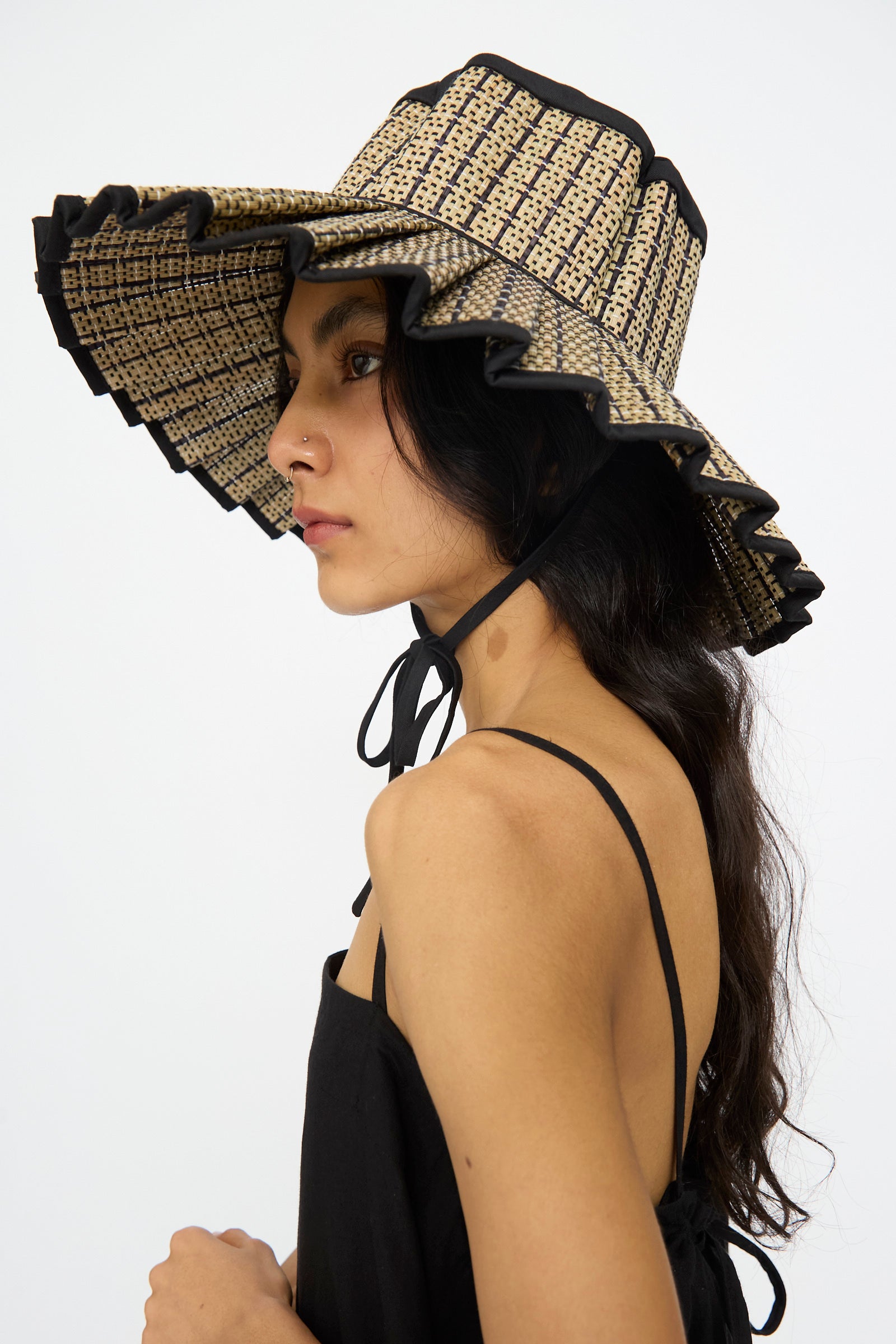 A person wears the Lorna Murray Island Capri Maxi Hat in Lovers Point, made from natural grass fibers with a black ruffle trim and chin ribbon, paired with a black spaghetti strap top.