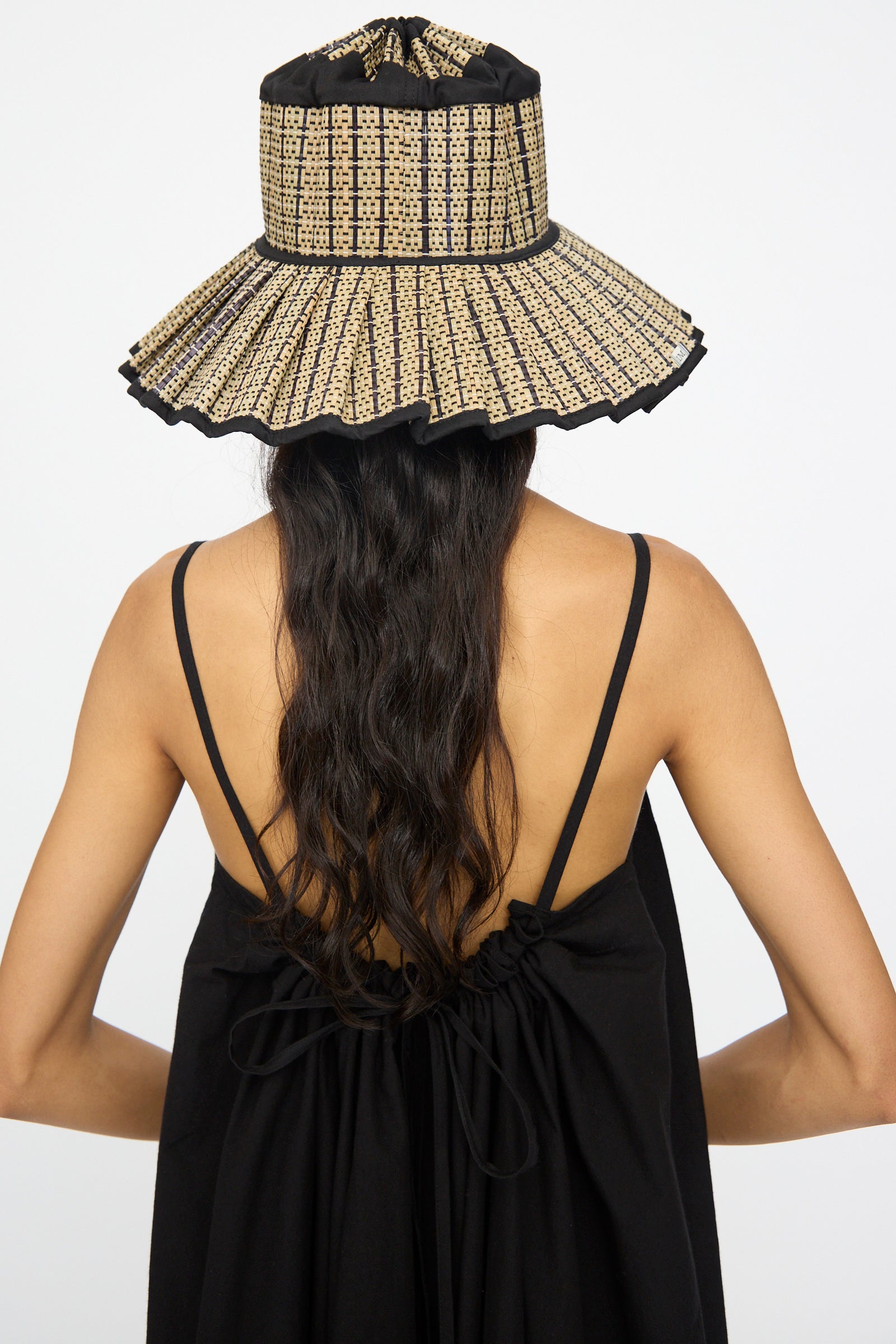 Person with long wavy hair in a black strappy dress wears the Lorna Murray Island Capri Maxi Hat in Lovers Point, a wide-brimmed sun hat, shown from the back against a plain background.