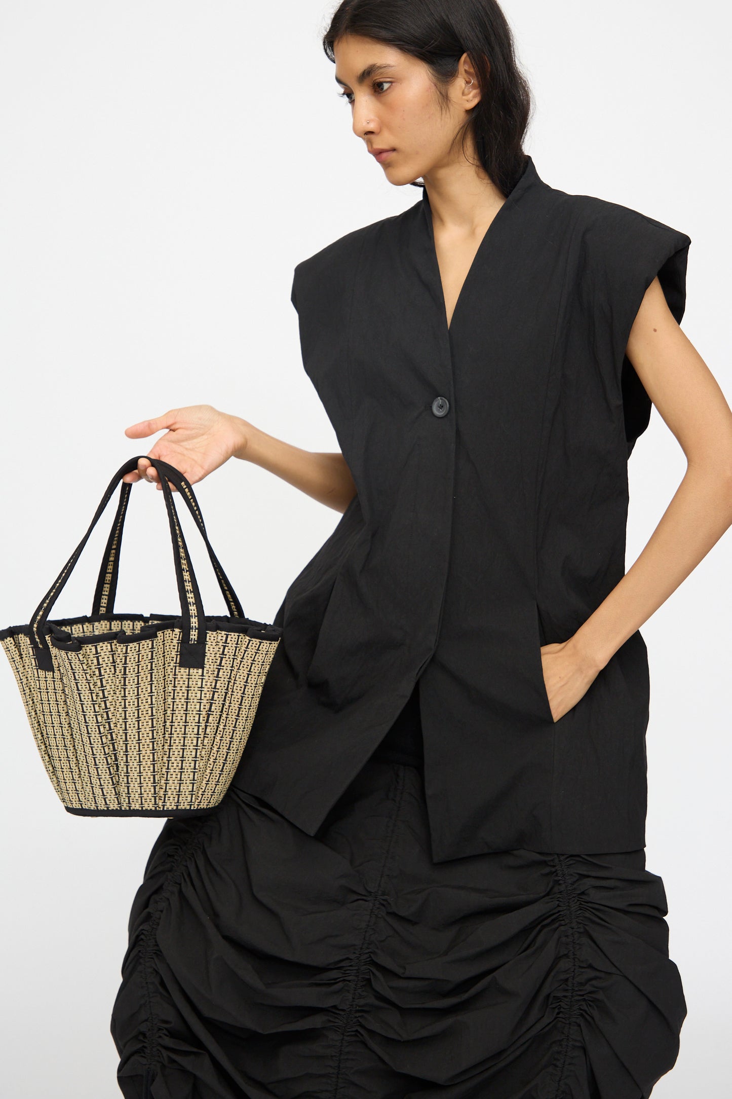A woman in a sleeveless black buttoned vest and ruched black skirt holds the Lorna Murray Mini Tropez Tote in Lovers Point, handmade in Indonesia from natural grass fibers, standing against a plain background.
