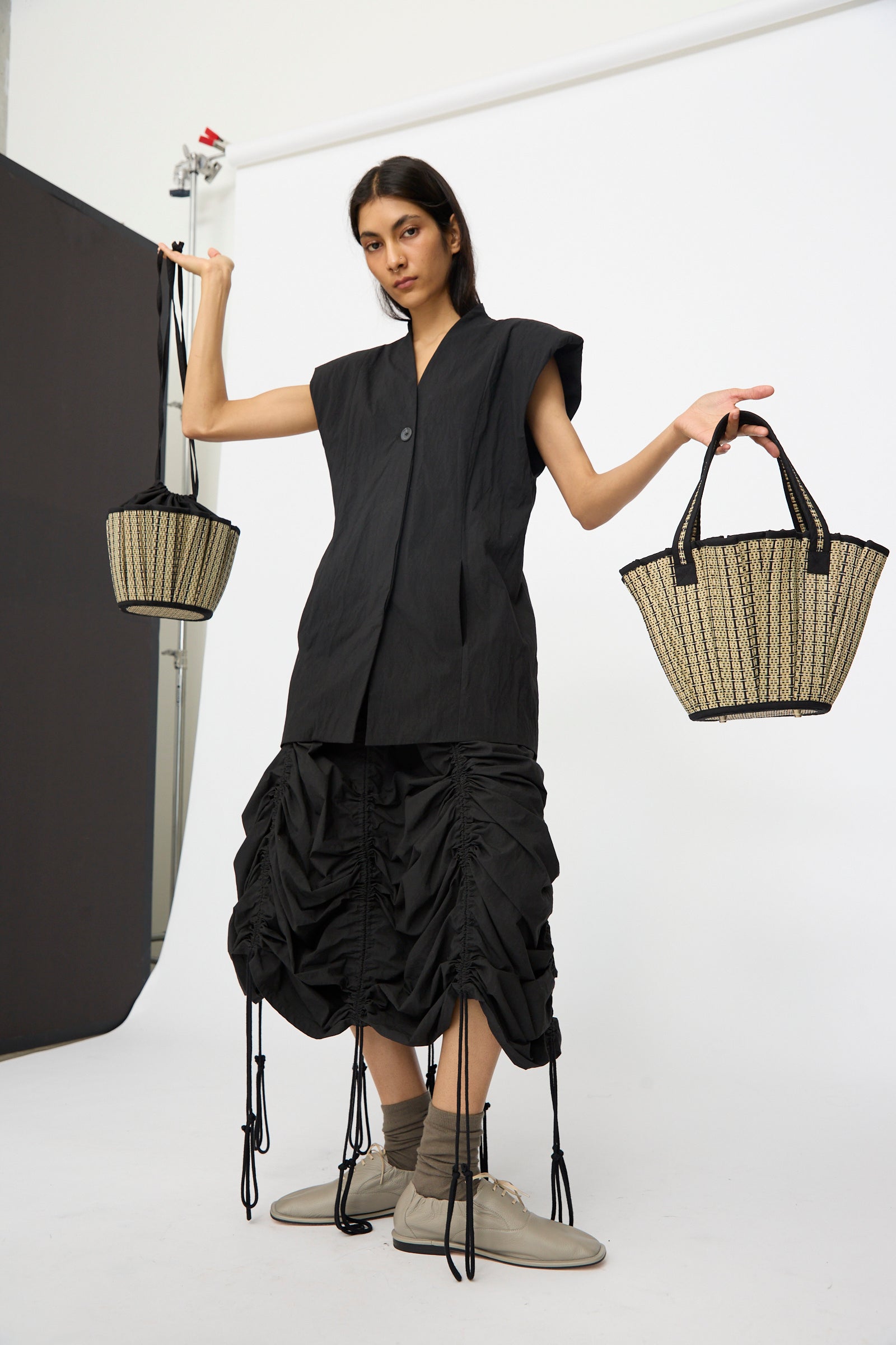 A person in a black sleeveless top and ruched skirt stands in a studio, raising both arms while holding two Lorna Murray Mini Tropez Tote bags in Lovers Point, each handmade in Indonesia.