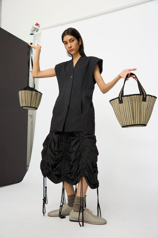 A person in a black sleeveless top and ruched skirt stands in a studio, raising both arms while holding two Lorna Murray Mini Tropez Tote bags in Lovers Point, each handmade in Indonesia.