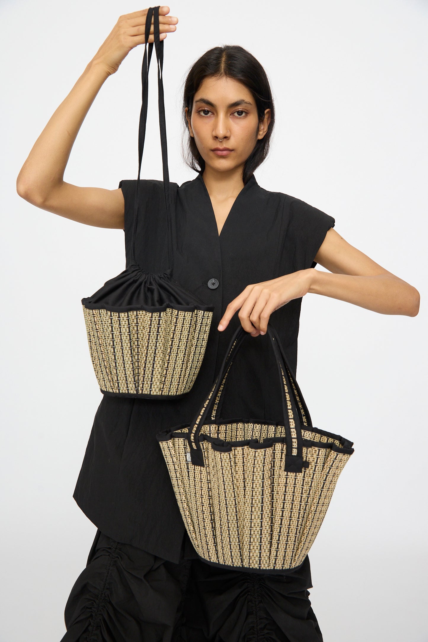 A person in a black outfit holds two Lorna Murray Mini Tropez Tote in Lovers Point straw handbags, each with black straps, one in each hand, handmade in Indonesia, against a plain white background.