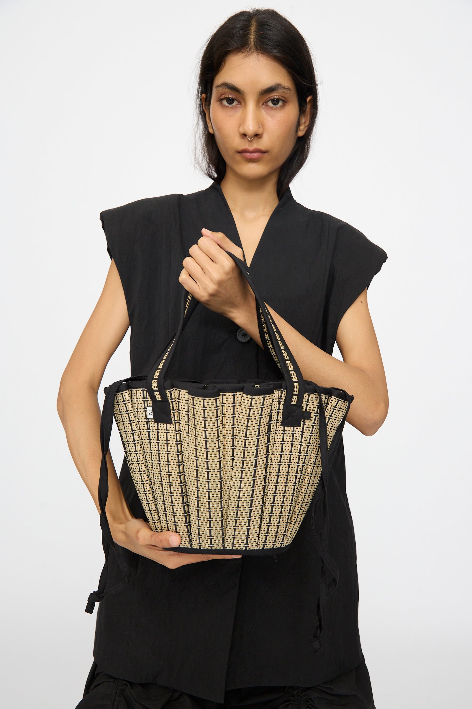 A person in a sleeveless black top holds the Lorna Murray Mini Tropez Tote in Lovers Point, handmade in Indonesia from natural grass fibers with beige and black patterns and two handles, set against a plain background.