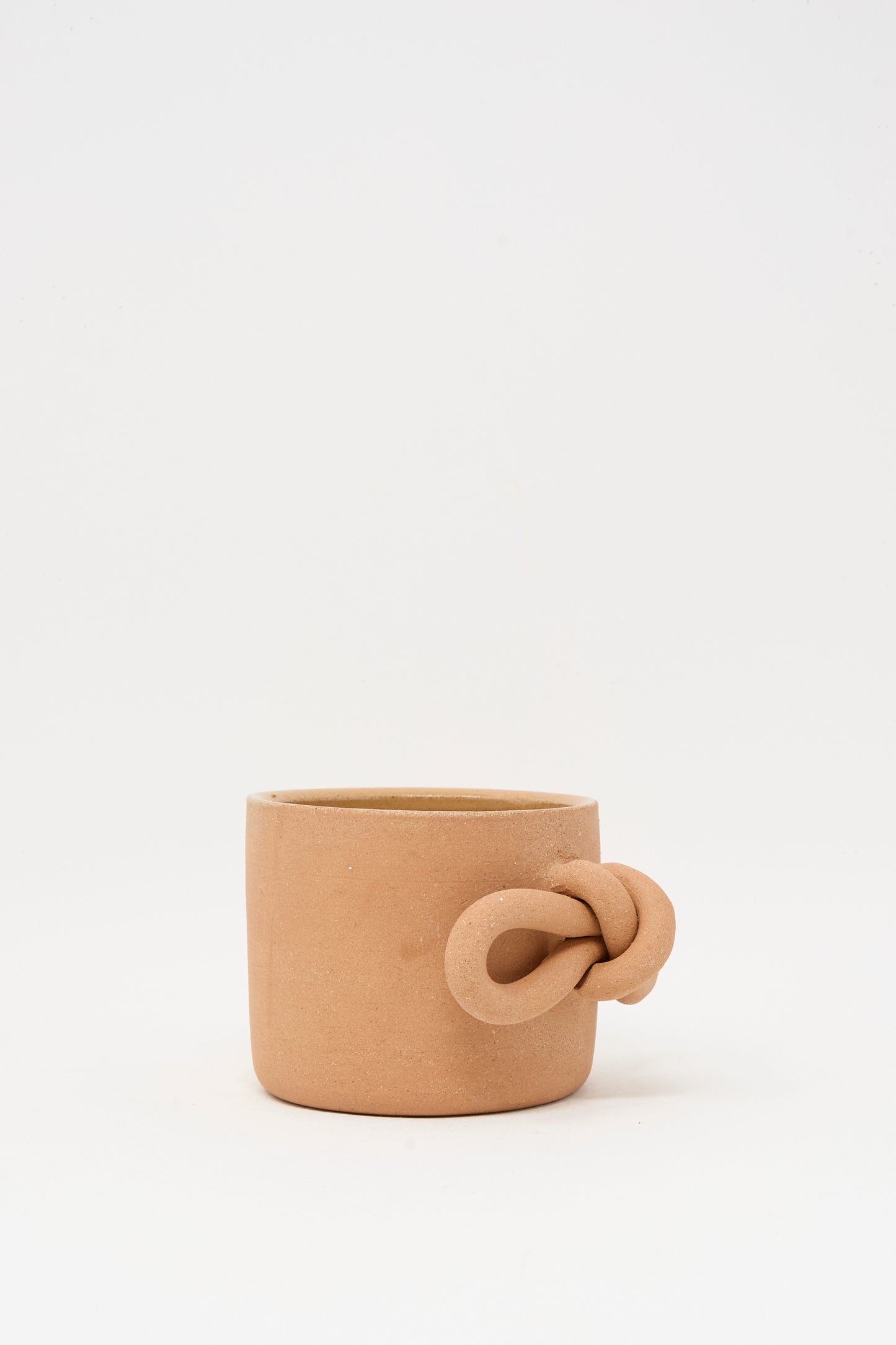 The Bow Knot Mug in Adobe by Lost Quarry features a plain, light brown ceramic body with a rounded shape and a unique knotted handle, exuding charm against a white background.