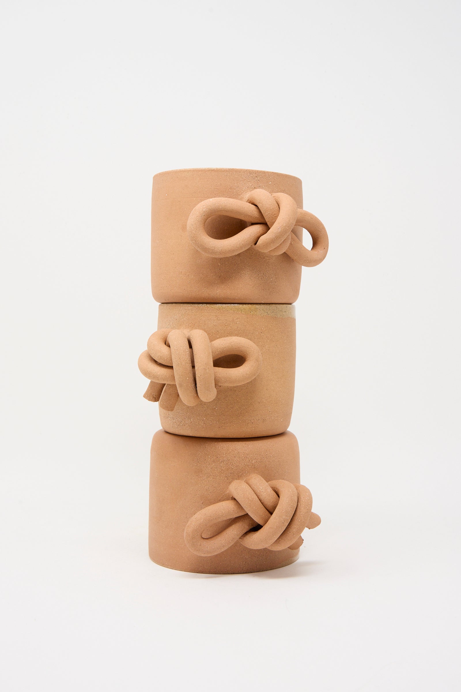 Three stacked Lost Quarry Bow Knot Mugs in Adobe, handcrafted from terracotta with a knot detail on the side, are shown against a white background—a striking display of artisanal design.