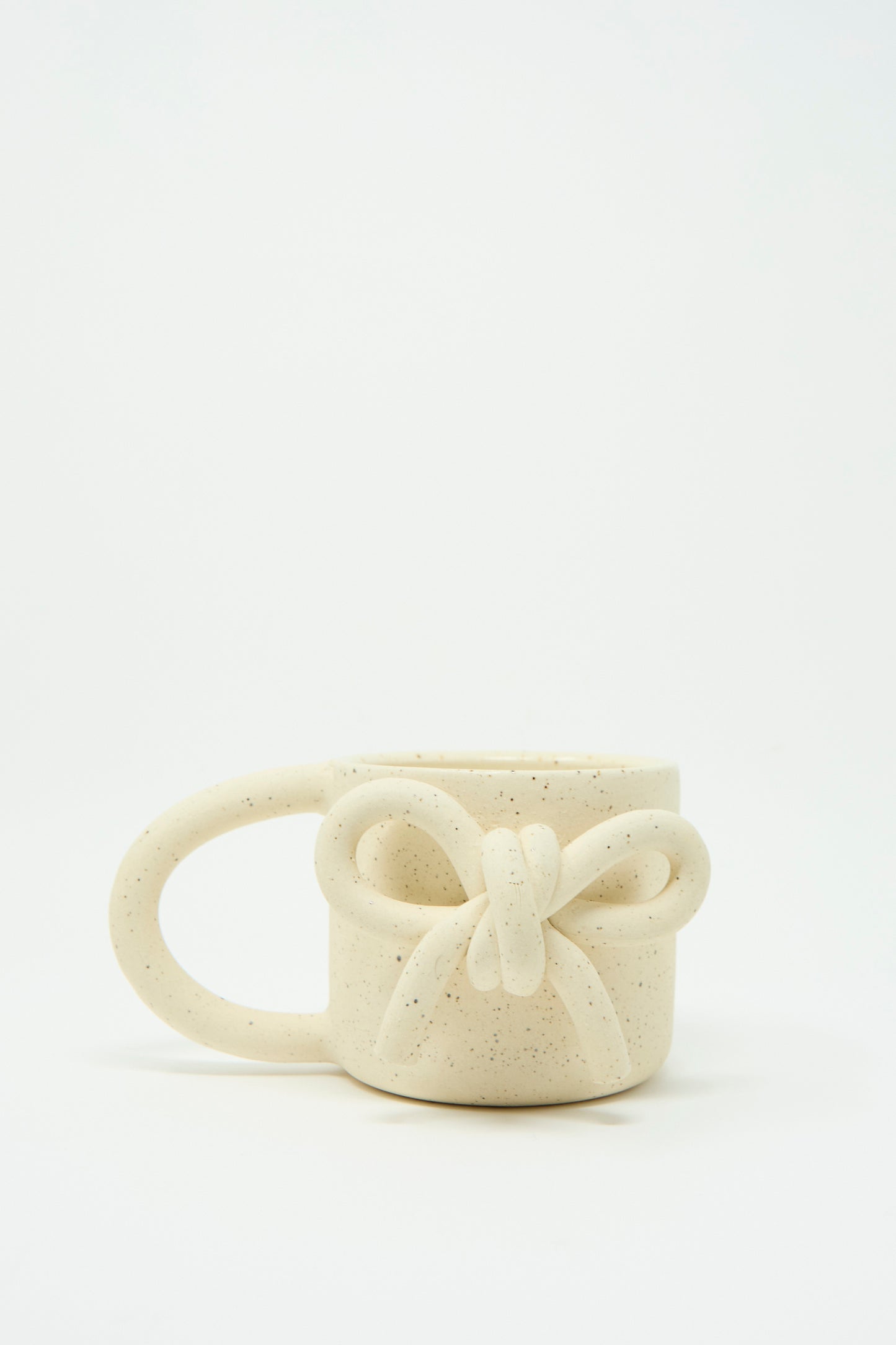 The Lost Quarry "Mug with Bow in Speckled White" features a distinctive oversized handle, with an intricately shaped bow at the front, against a white background. 