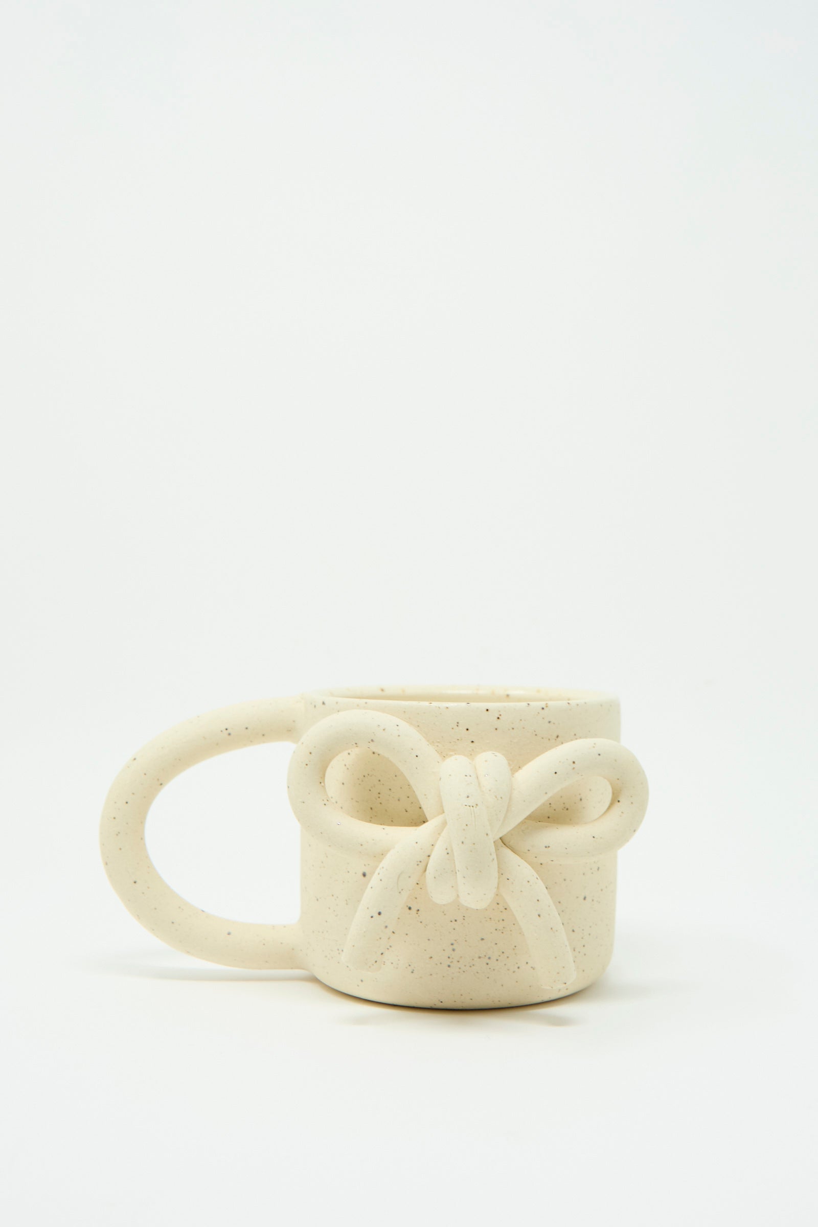 The Lost Quarry "Mug with Bow in Speckled White" features a distinctive oversized handle, with an intricately shaped bow at the front, against a white background. 