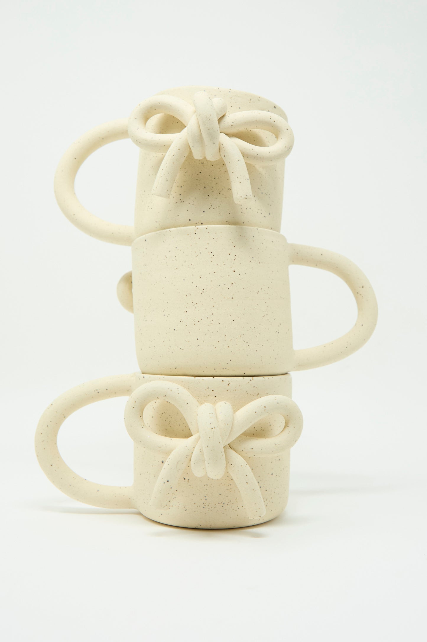 A trio of Lost Quarry's Mug with Bow in Speckled White, each showcasing an oversized handle and a twisted bow at the front, is stacked vertically against a pristine white background.