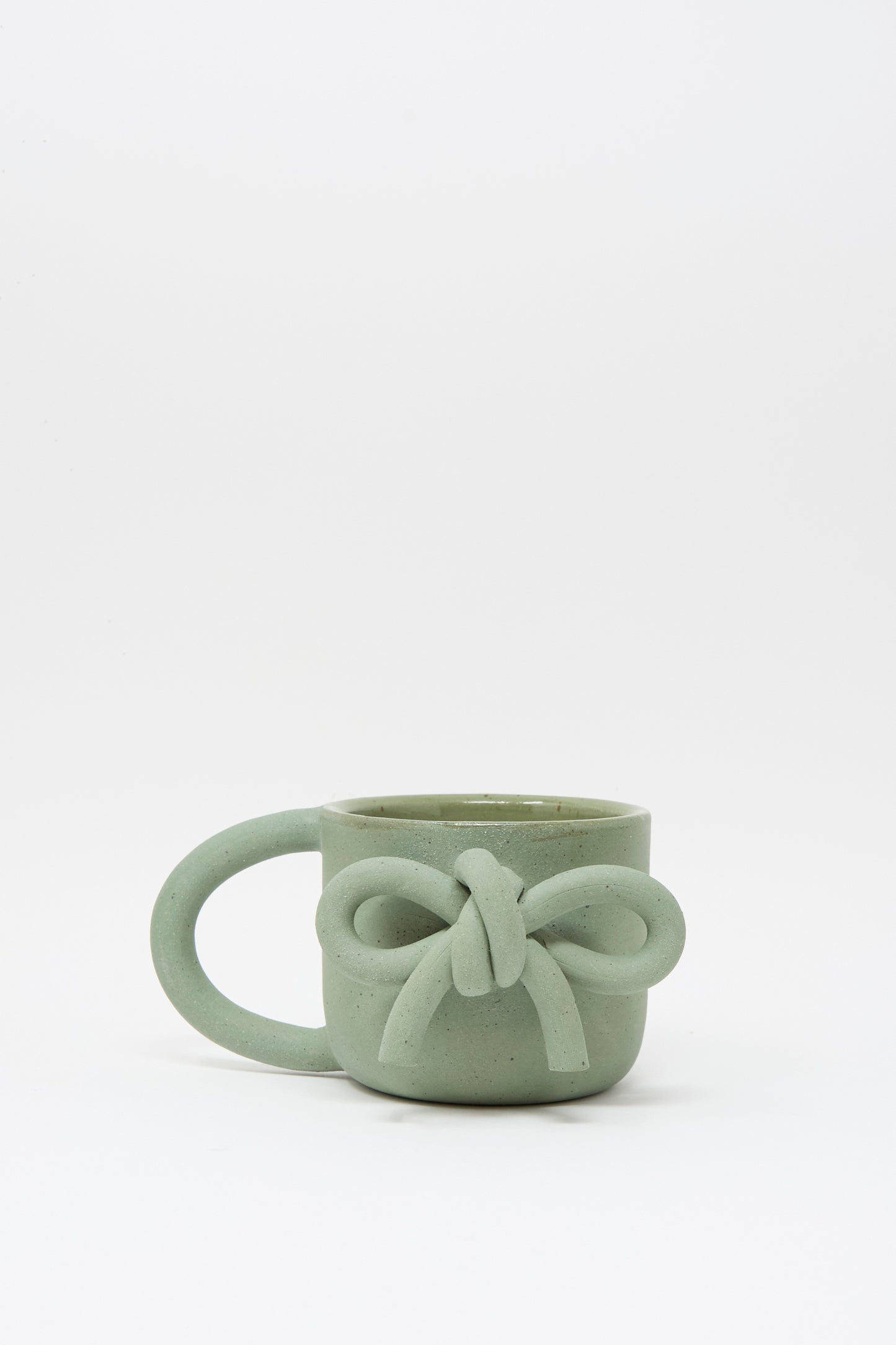 The Lost Quarry Hand Built Bow Mug in Sage features a light green ceramic finish with a decorative bow, an oversized handle, and is shown against a white background.