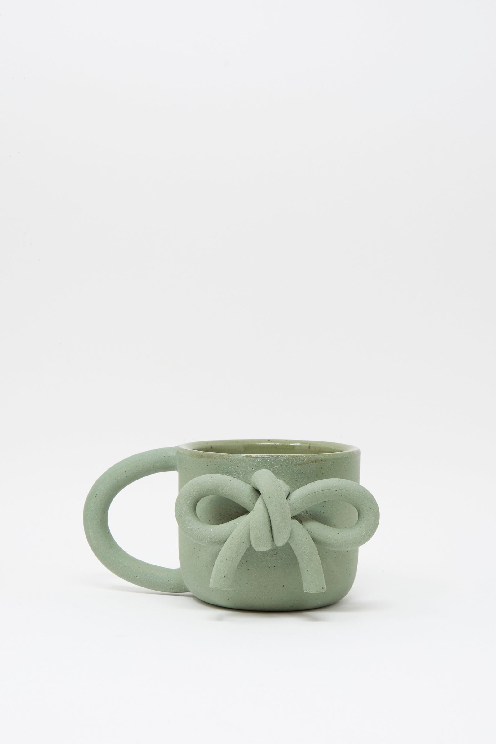 The Lost Quarry Hand Built Bow Mug in Sage features a light green ceramic finish with a decorative bow, an oversized handle, and is shown against a white background.