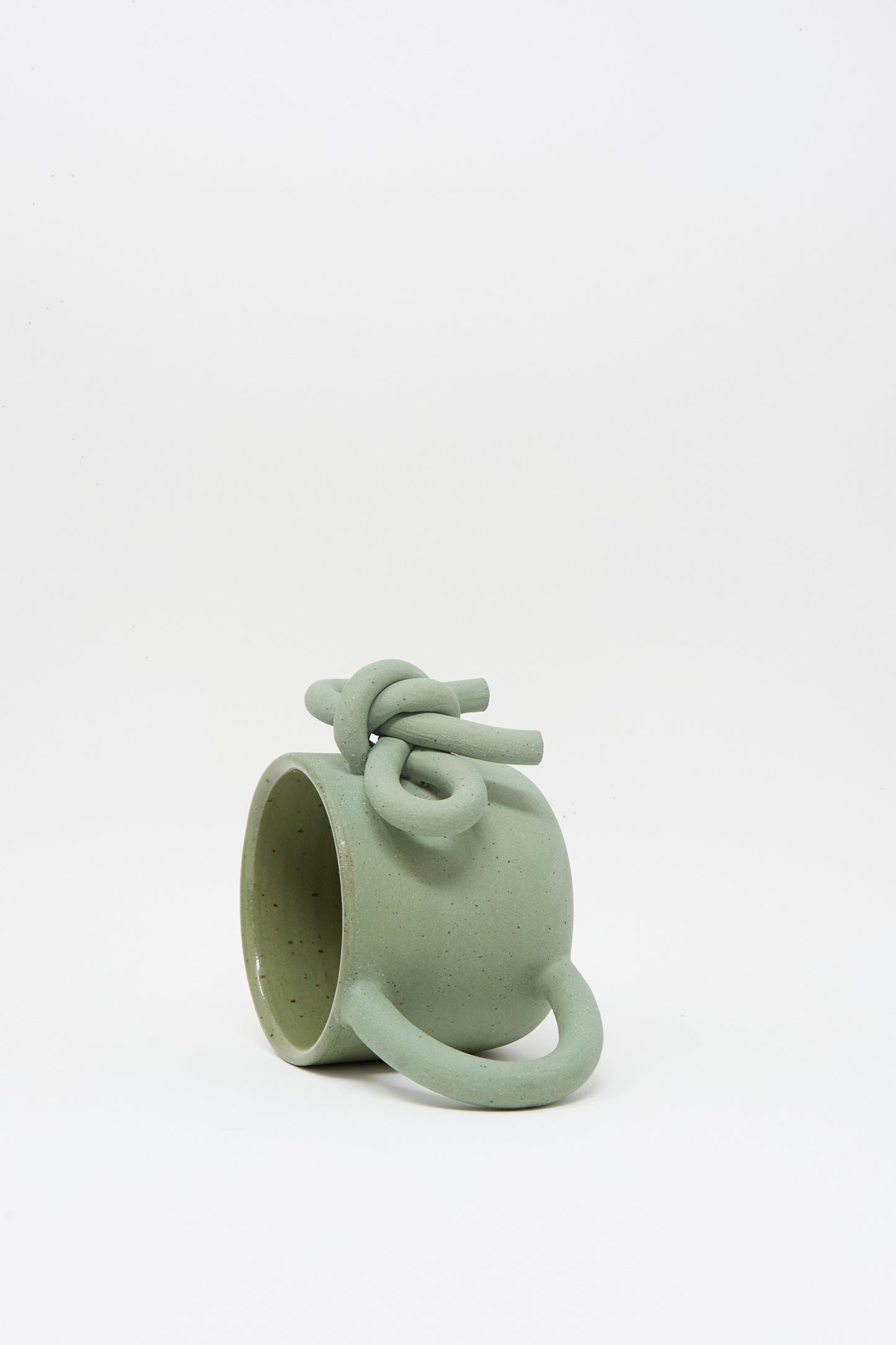 The Lost Quarry Hand Built Bow Mug in Sage features a light green ceramic body with a twisted knot design on the rim, an oversized handle, and a soft sage glaze, displayed against a plain white background.