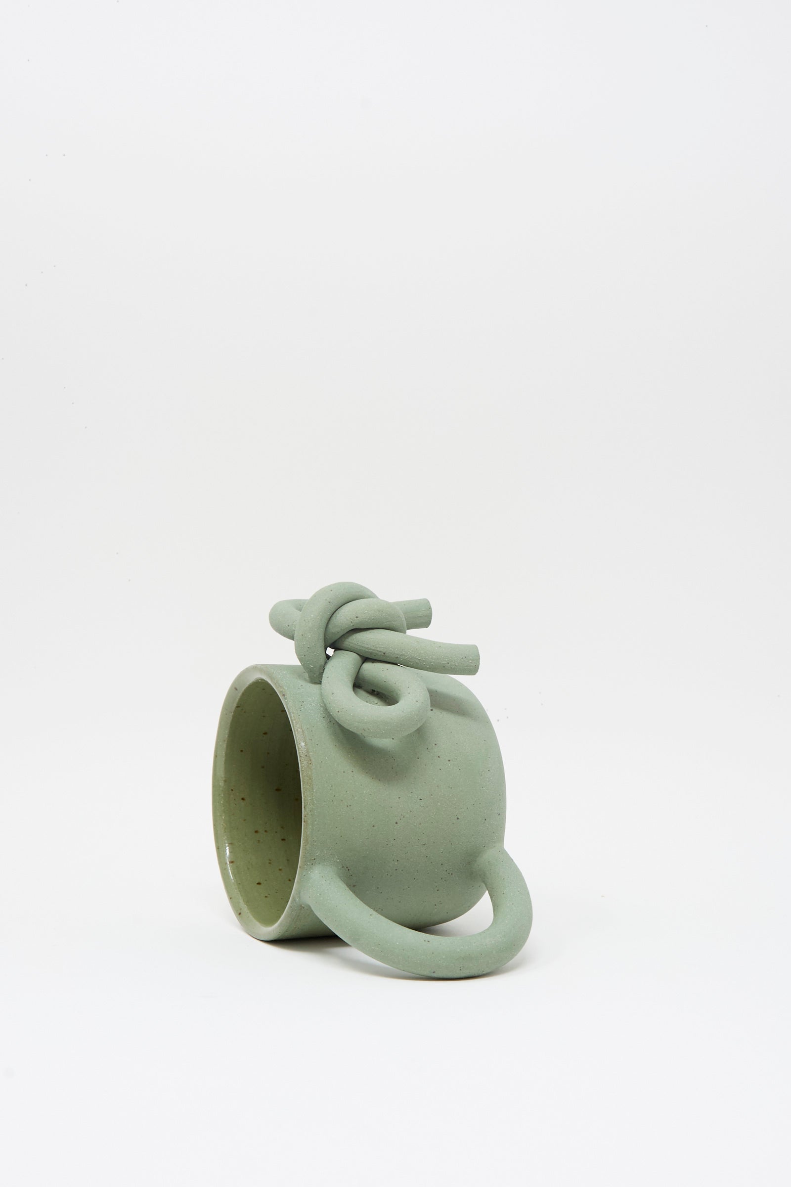 The Lost Quarry Hand Built Bow Mug in Sage features a light green ceramic body with a twisted knot design on the rim, an oversized handle, and a soft sage glaze, displayed against a plain white background.