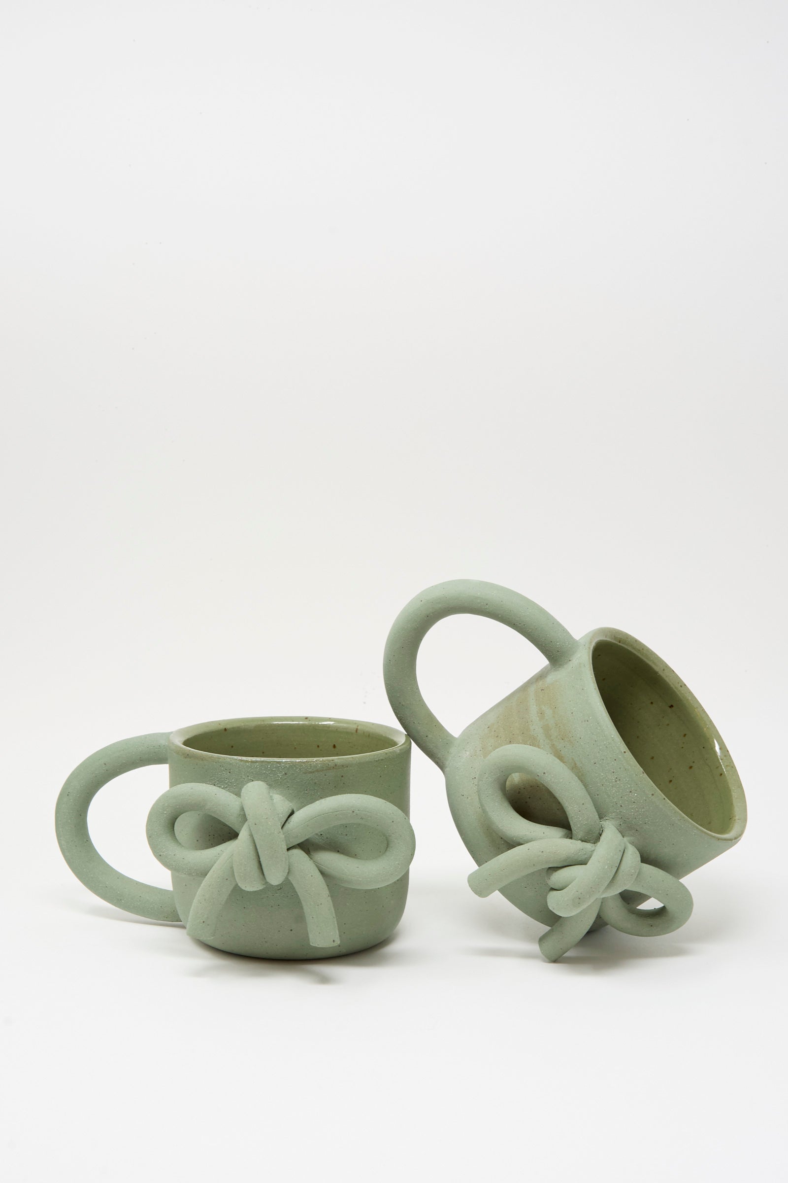 The Hand Built Bow Mugs in Sage by Lost Quarry are two light green ceramic mugs with oversized handles and bow-shaped accents, featuring a smooth sage glaze, shown against a plain white background.